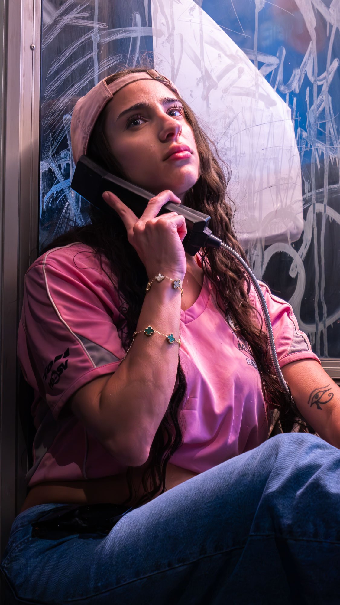 Young woman with long brown hair, wearing a pink jersey and a beige cap, sitting in a phone booth with graffiti-covered glass. She is holding a vintage black telephone receiver to her ear and looking thoughtfully into the distance.