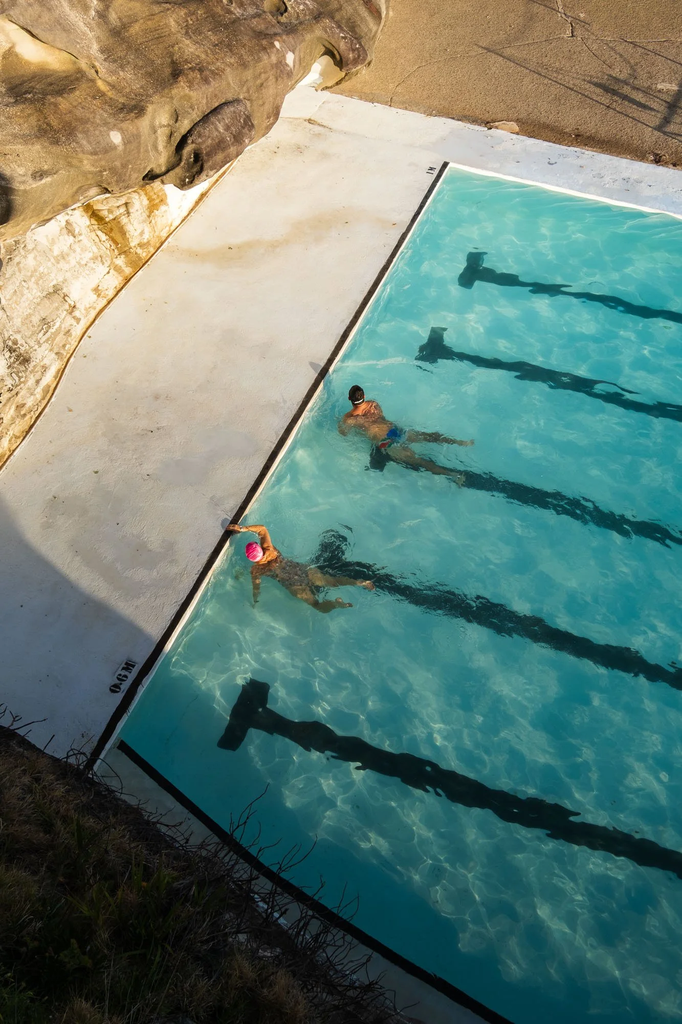 A person swimming in an outdoor swimming pool next to a rocky wall and a sandy area, with a towel or clothing hanging on the edge of the pool.