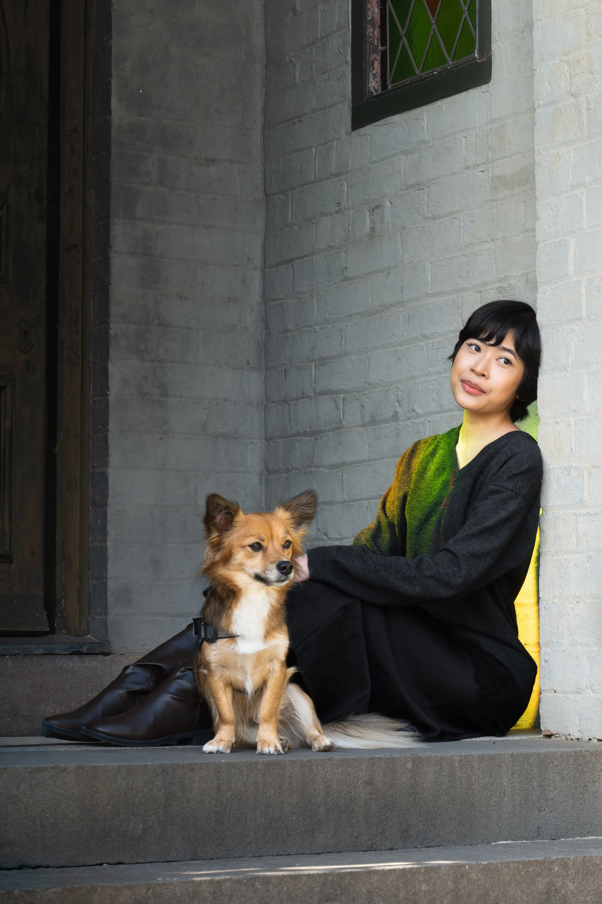 A woman with short black hair wearing a black and yellow sweater, sitting on steps outside near a white brick wall, holding a tan and white dog with a black harness, and a brown puppy sitting on the steps in front of her.