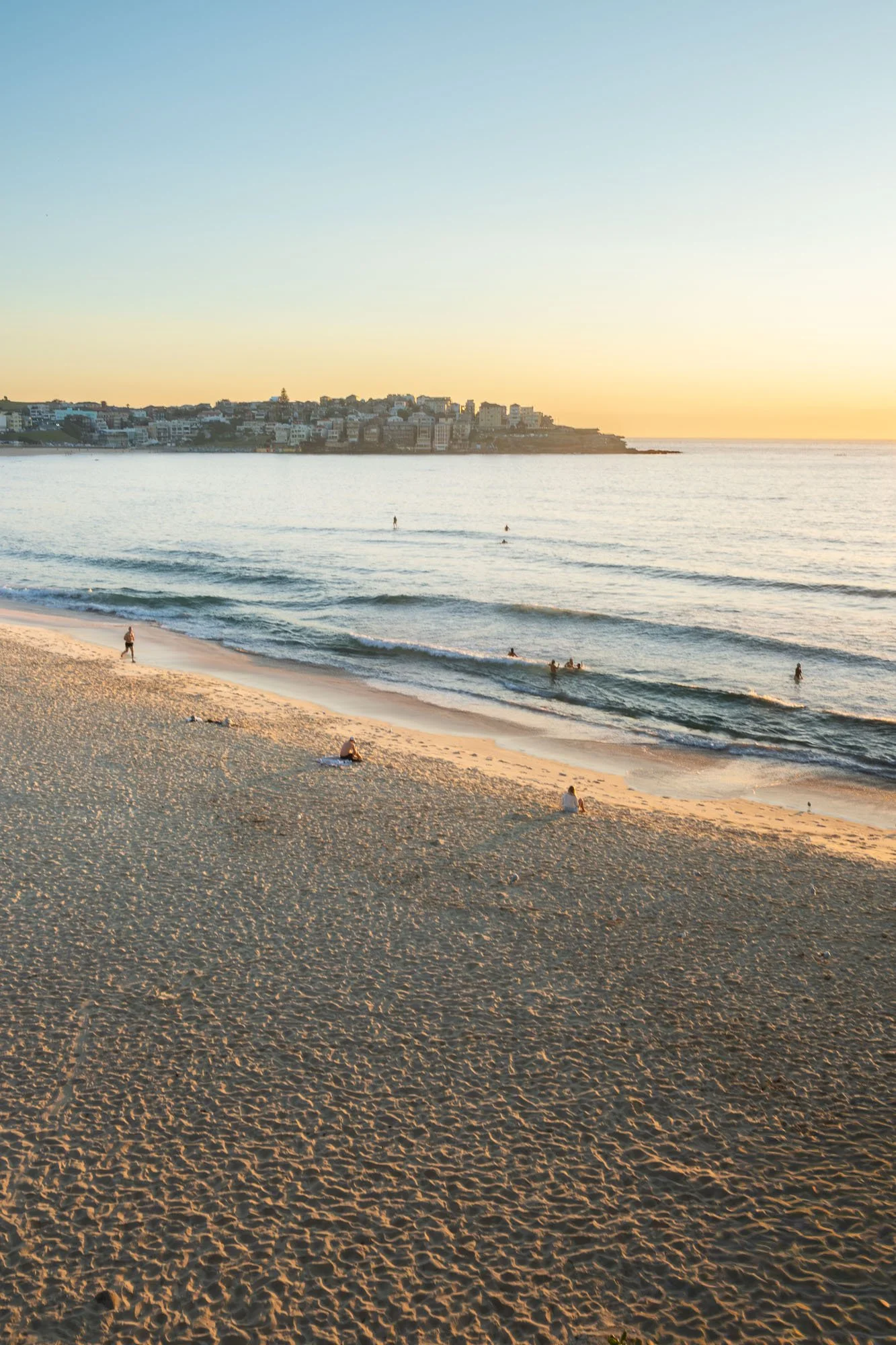 Sunset over a sandy beach with a few people walking and sitting, with a town on a hill in the background.