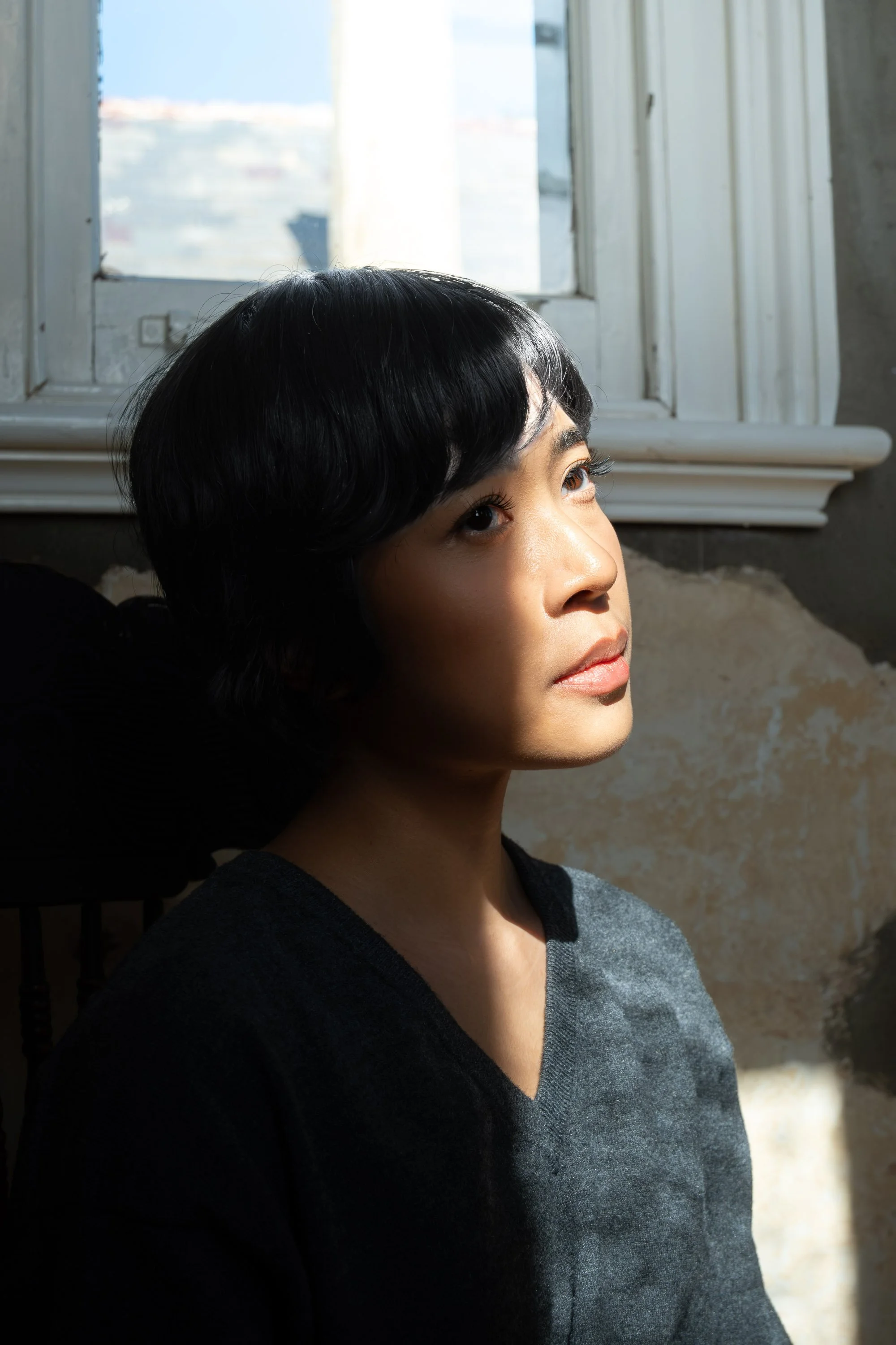 A young woman with short black hair and light brown skin gazes to her right, standing next to a window with sunlight illuminating her face and black top.