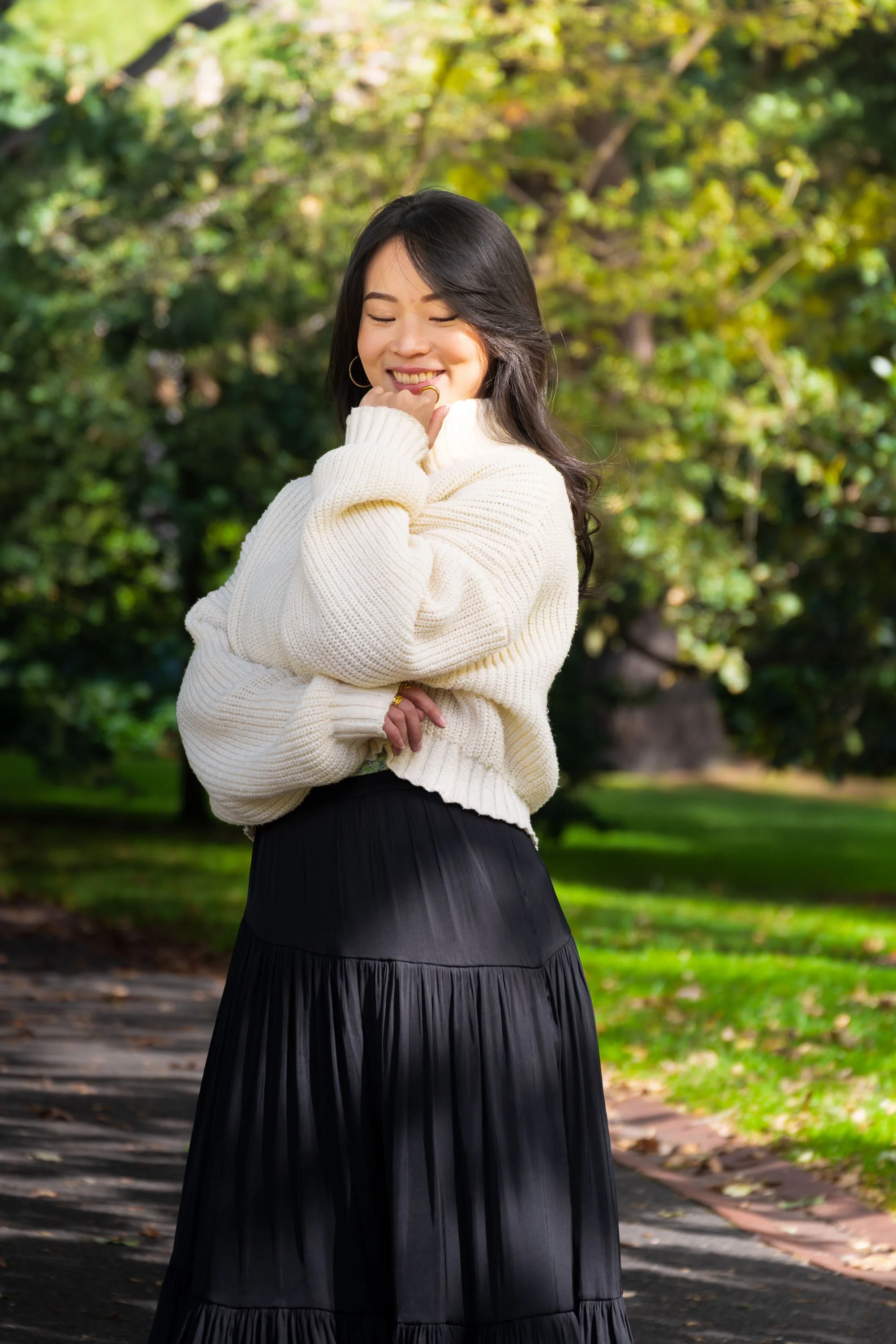 A young woman with black hair, wearing a cream-colored sweater, smiling with her eyes closed, standing outdoors on a wooden pathway in a park with green trees in the background.