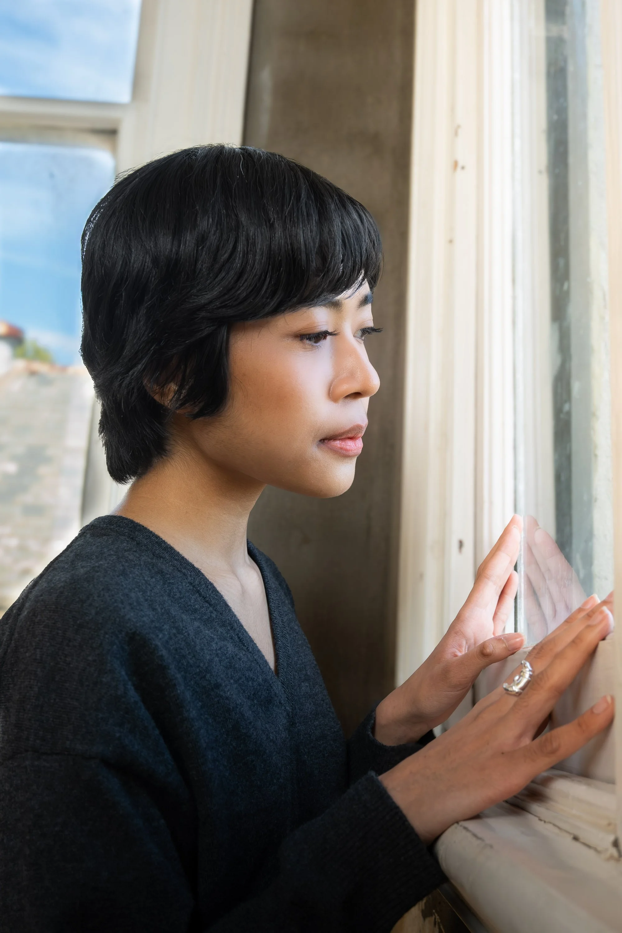 A woman with short black hair wearing a black sweater touching a window with her hands.