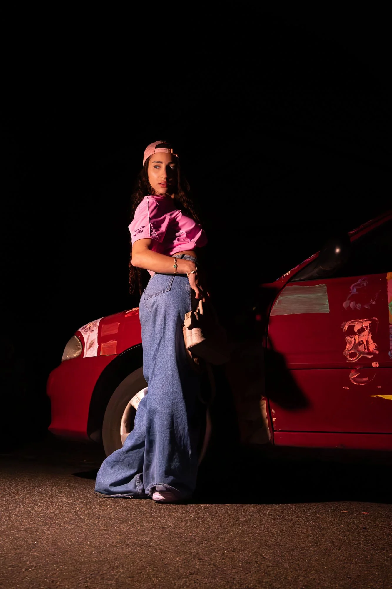 Woman in pink cap and pink top leaning on red car at night with dark sky.