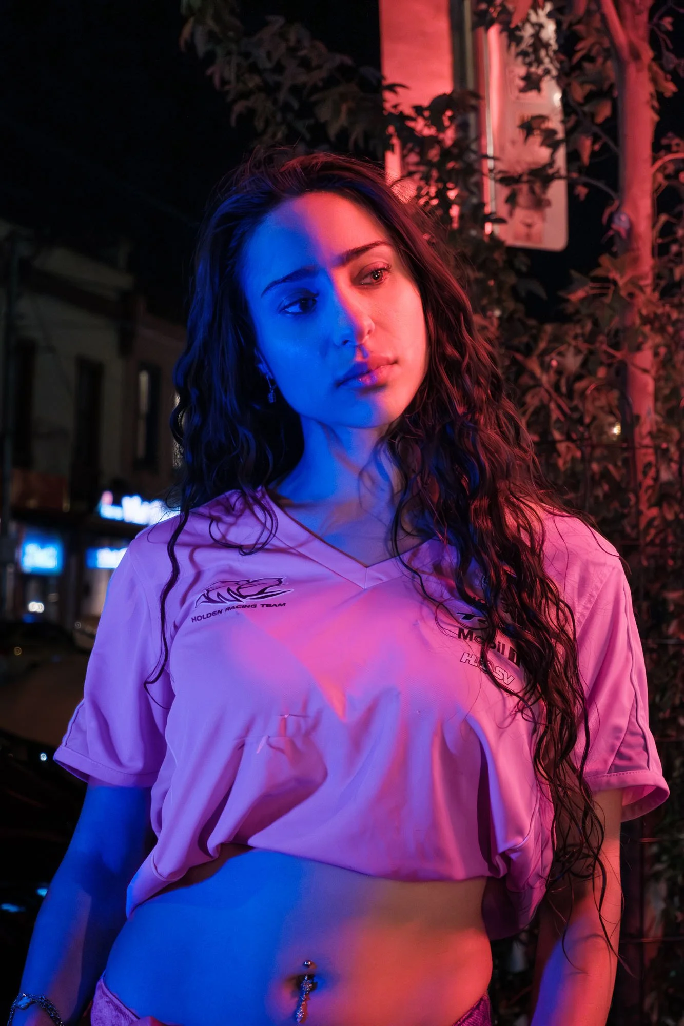 A young woman with long wavy hair stands outdoors at night illuminated by pink and blue neon lights. She wears a racing team shirt and has a belly piercing. In the background, blurred city lights and trees are visible.