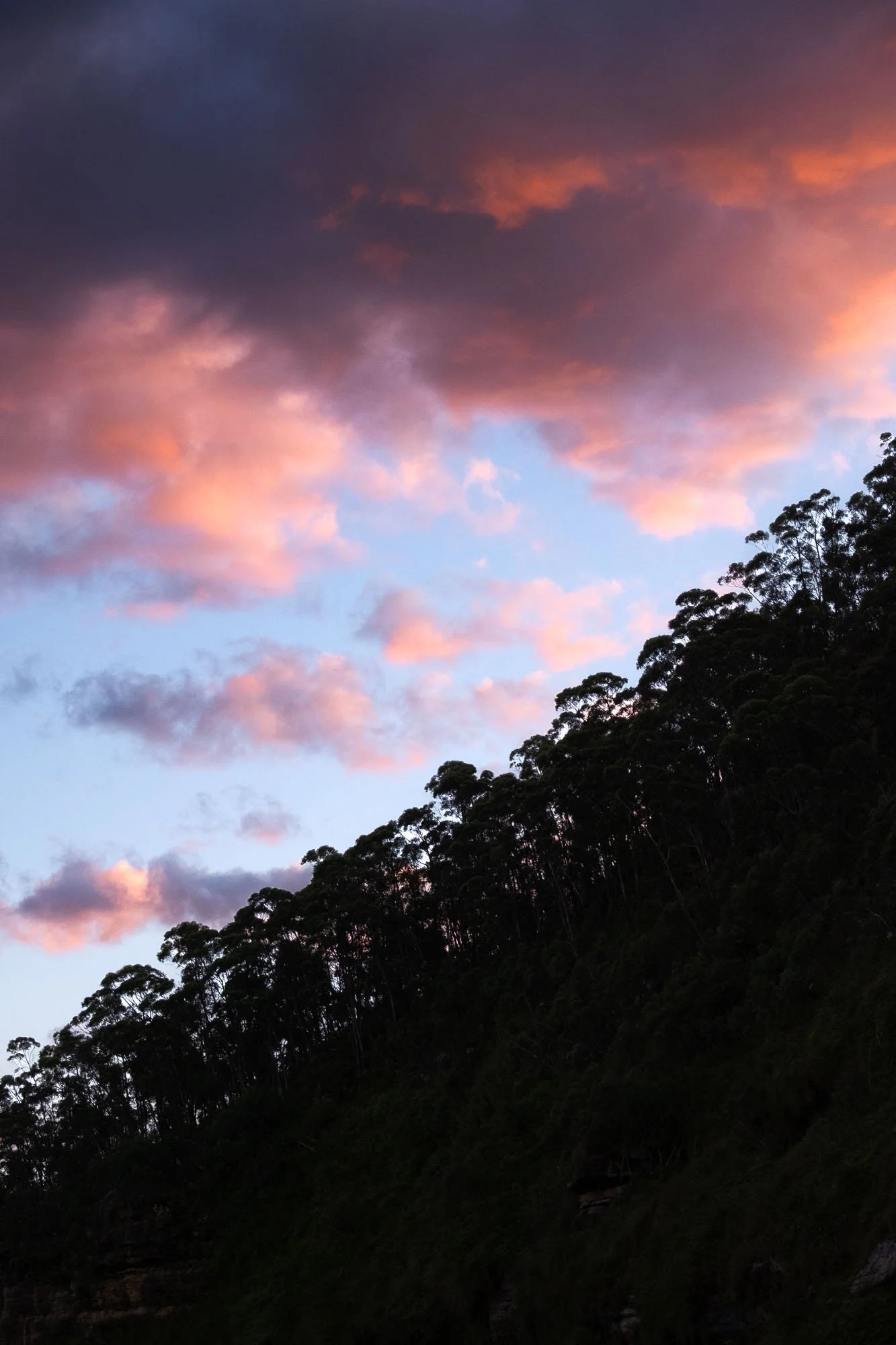 Sunset sky with pink and purple clouds above a silhouette of a forested hillside.