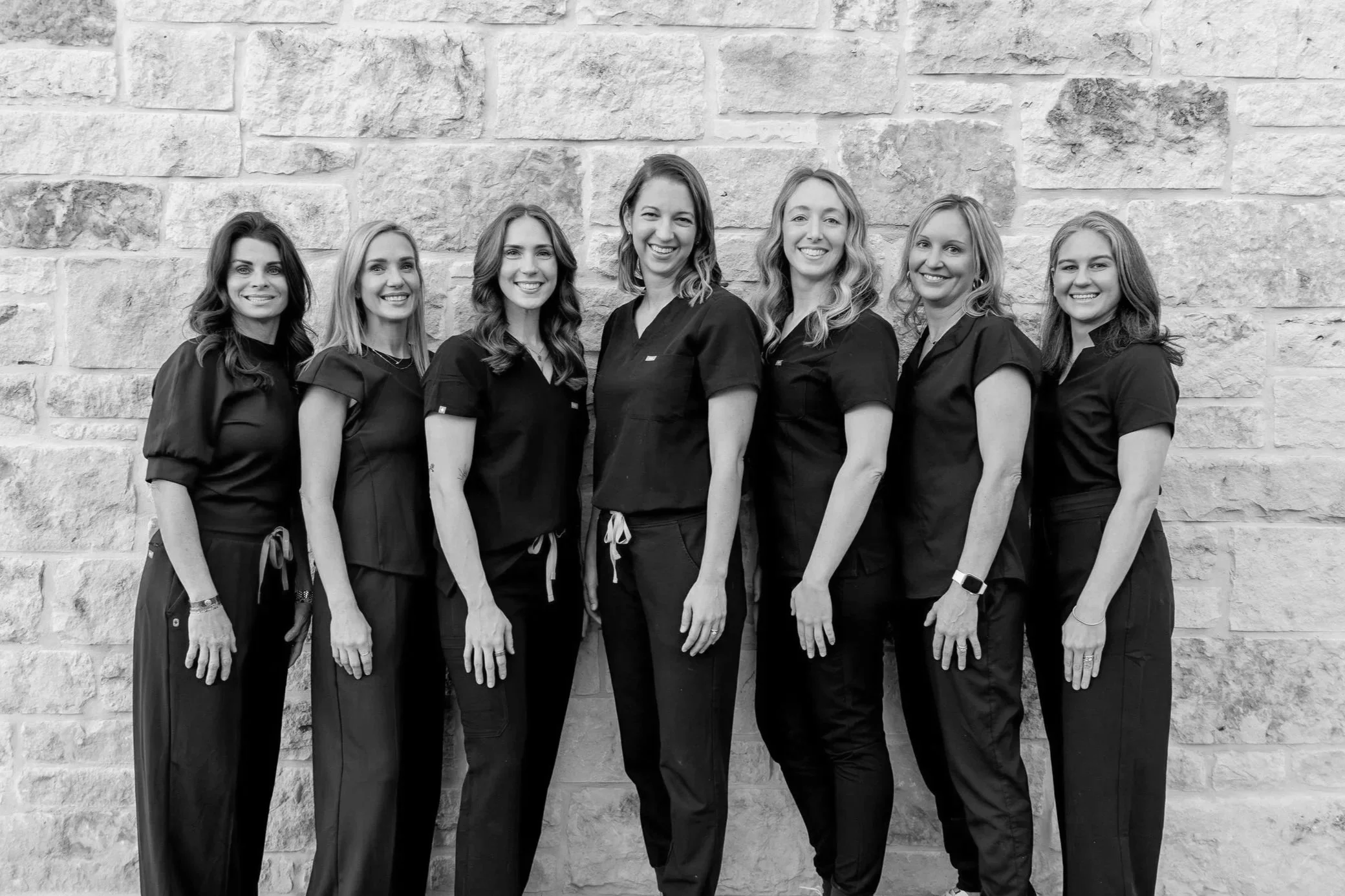 Seven women standing side by side in front of a stone wall, wearing black scrubs and smiling.