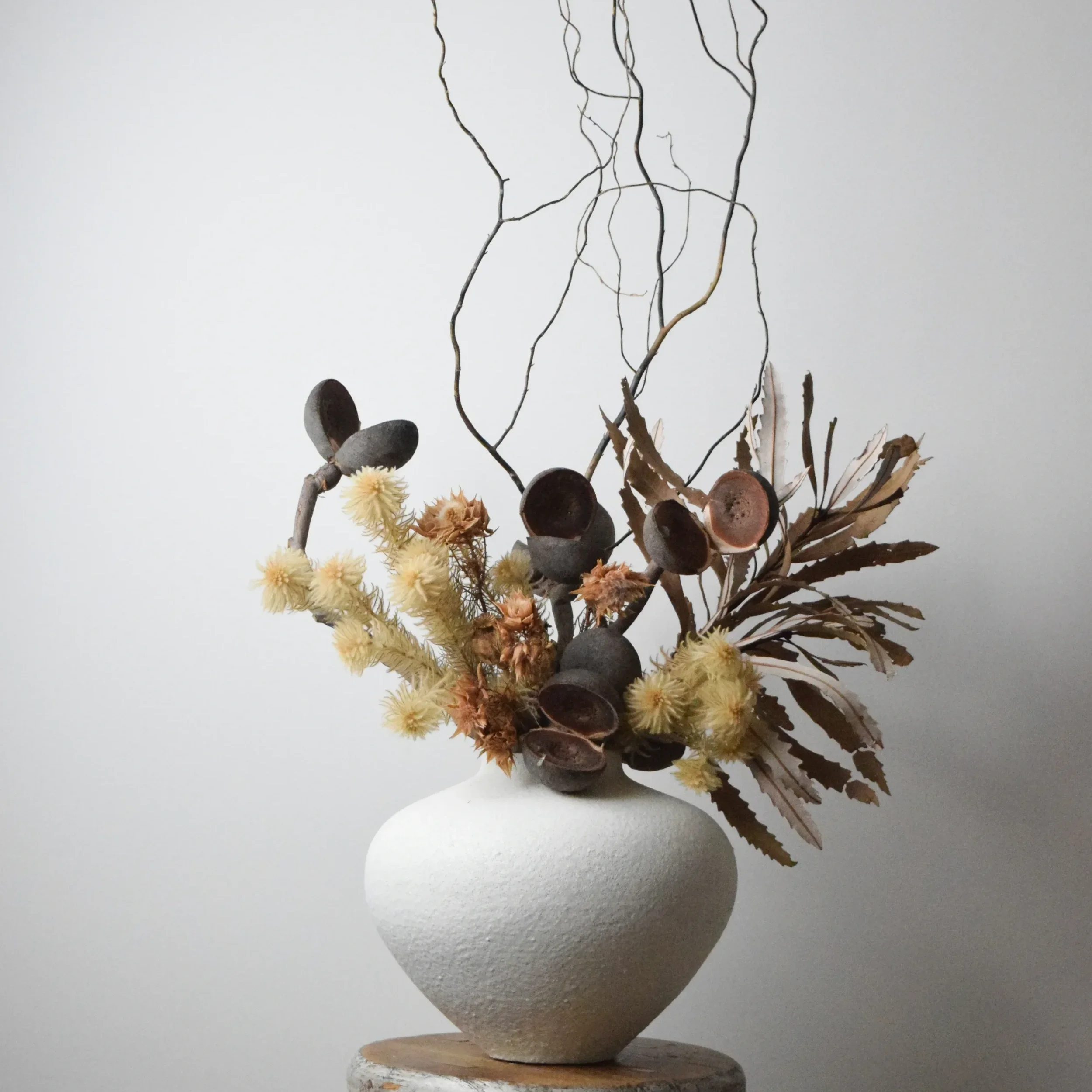 A minimalist art installation featuring a white pedestal with a flat, round top, supporting a clear glass or acrylic container filled with various natural elements like dry branches, leaves, flowers, and small black stones, set against a plain gray background.