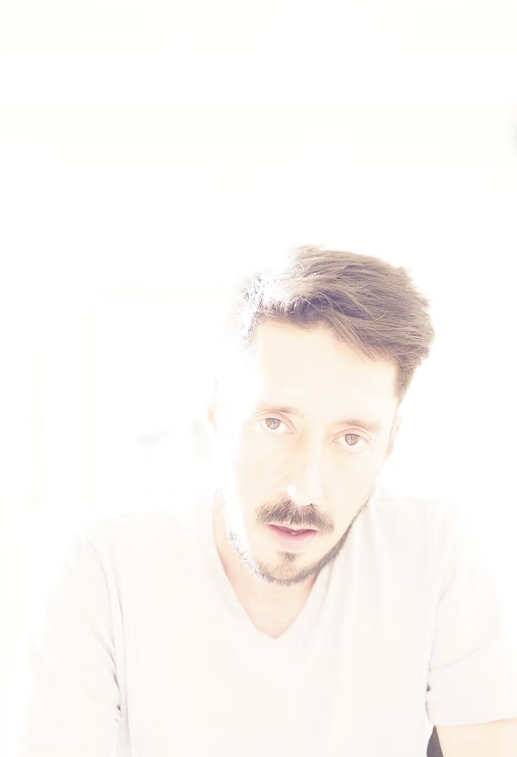 A man with dark hair and a mustache, wearing a light-colored shirt, sitting against a bright, overexposed background.
