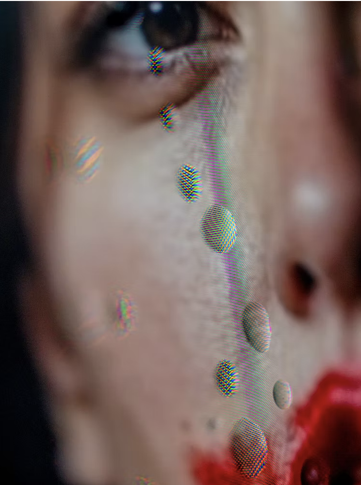 Close-up of a woman's face with makeup, with colorful droplets or beads running down her cheek, and red lipstick.