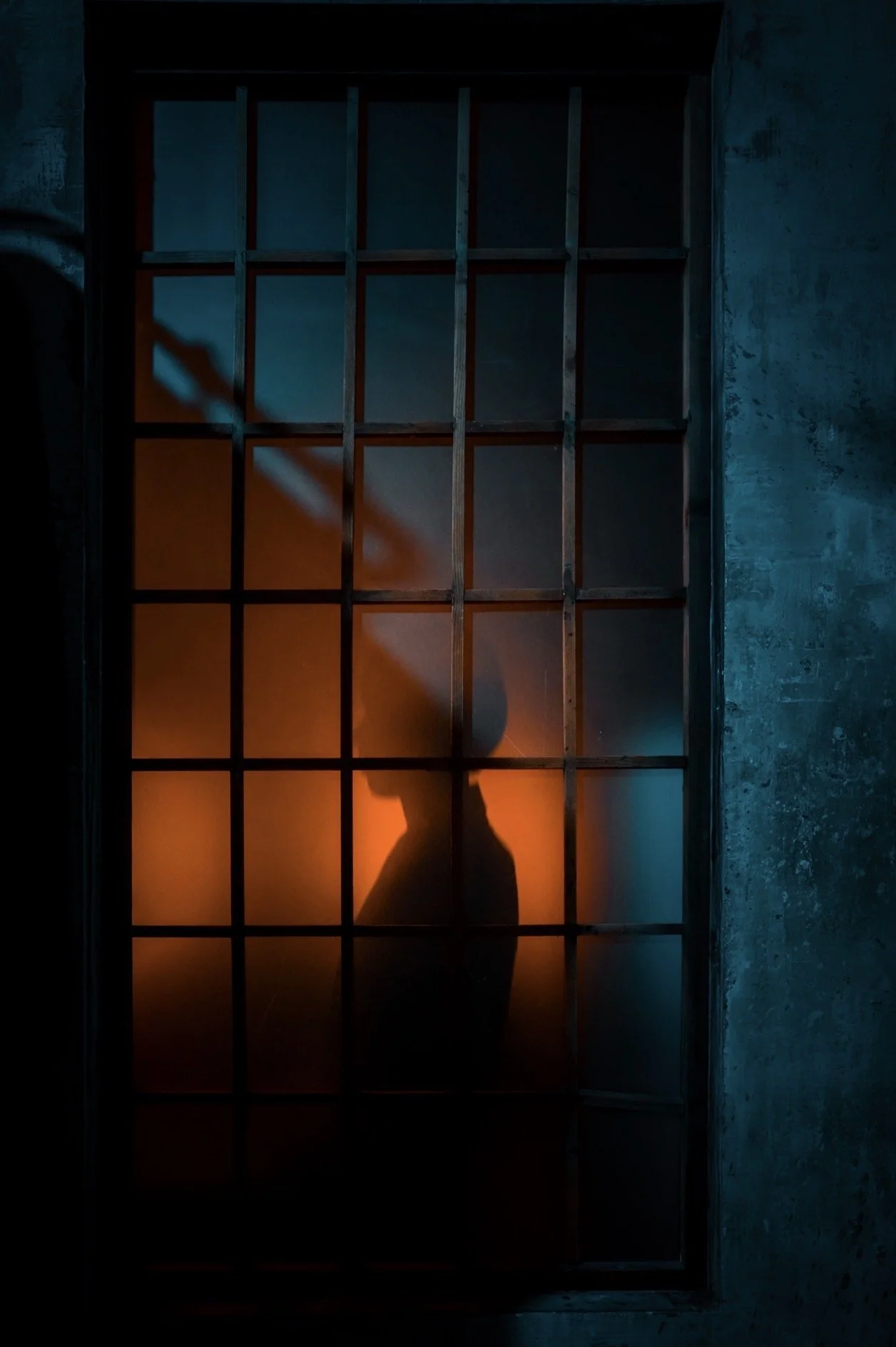 Silhouette of a person wearing a hat, standing behind a barred window with dim blue and orange lighting
