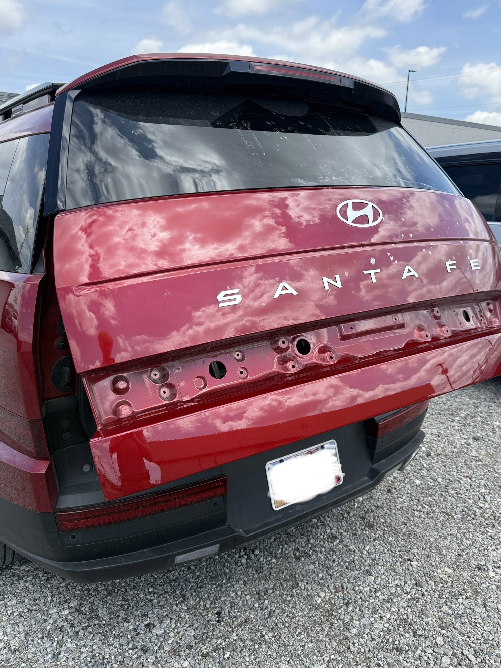 Rear view of a red Hyundai Santa Fe SUV with the back hatch open, showing the vehicle's interior and missing rear tailgate, parked on gravel with buildings and sky in the background.
