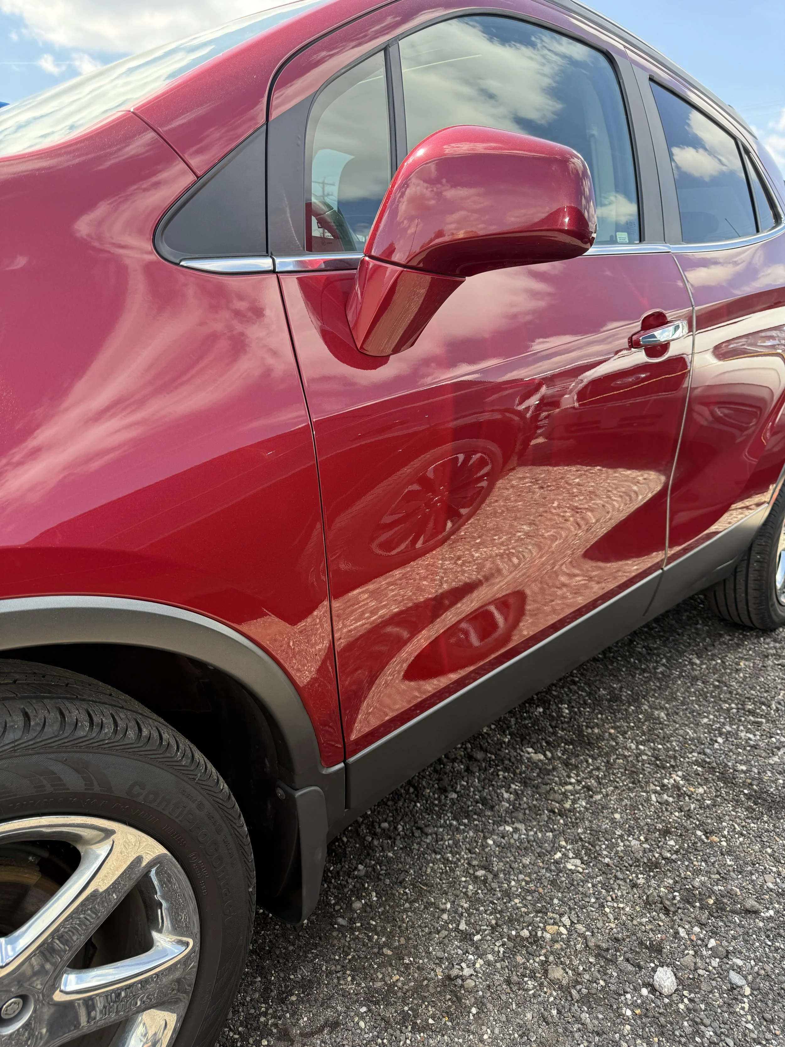 Close-up of a red SUV parked on a gravel lot, showing the front wheel, side mirror, and a portion of the driver's side window, with reflections of the sky and clouds in the window.