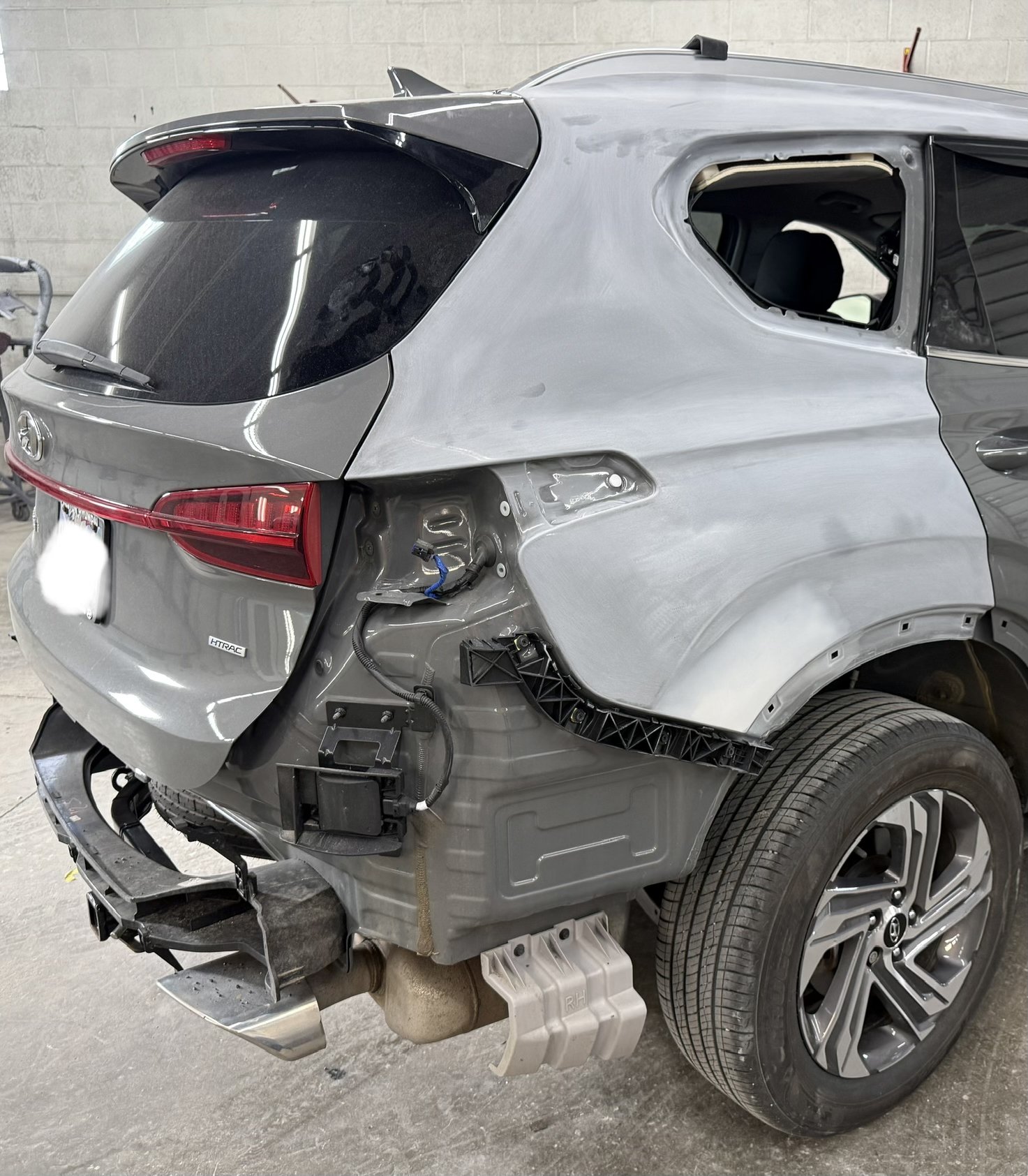 Partially disassembled gray car in a workshop, missing rear bumper and some panels, with exposed wiring and components, and a visible rear tire.