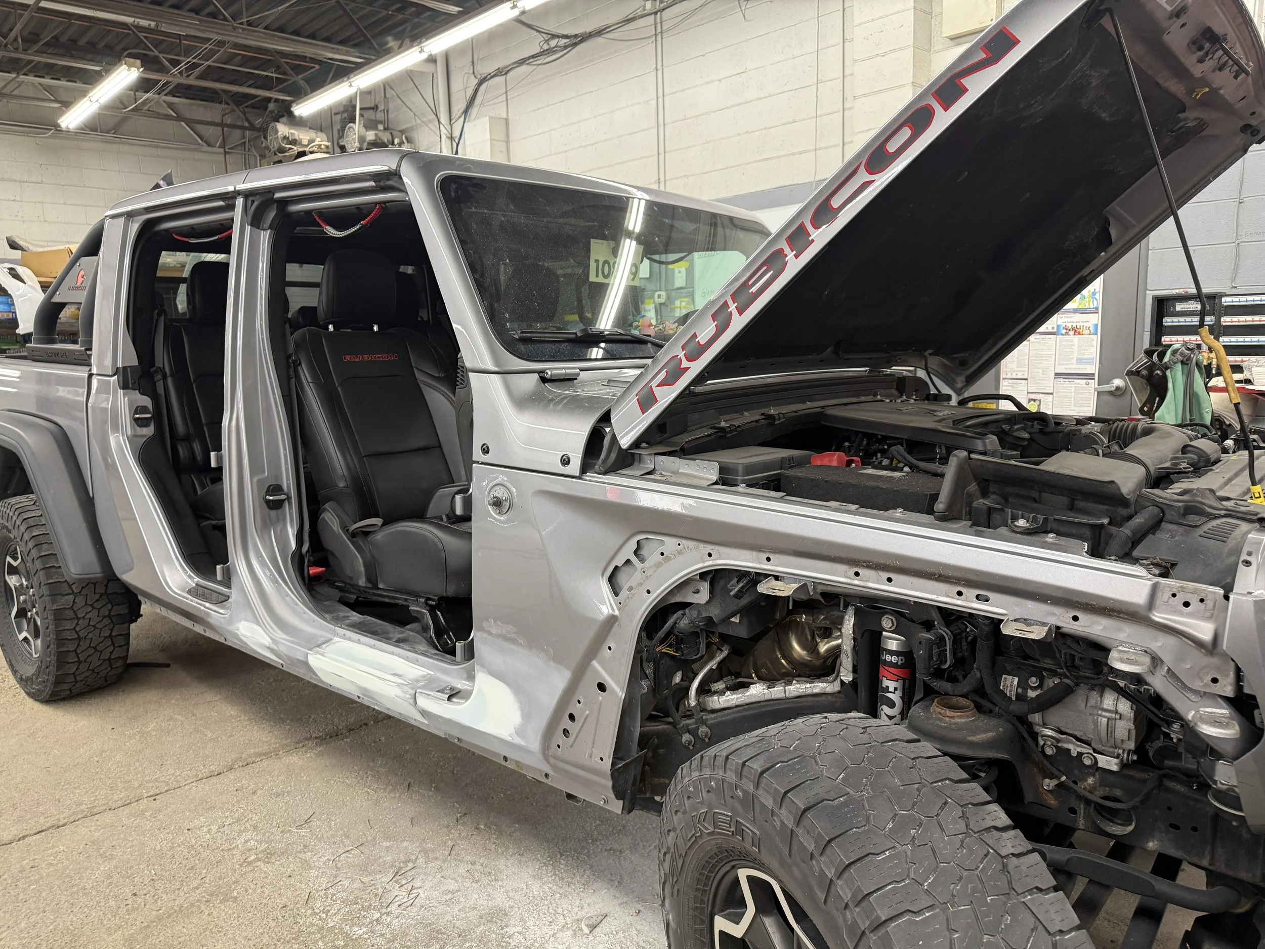 A silver Jeep Gladiator truck inside a garage with its hood open, revealing the engine. The vehicle is missing its front grille and part of the front bumper. The garage has fluorescent lighting, white brick walls, and various tools hanging or placed 