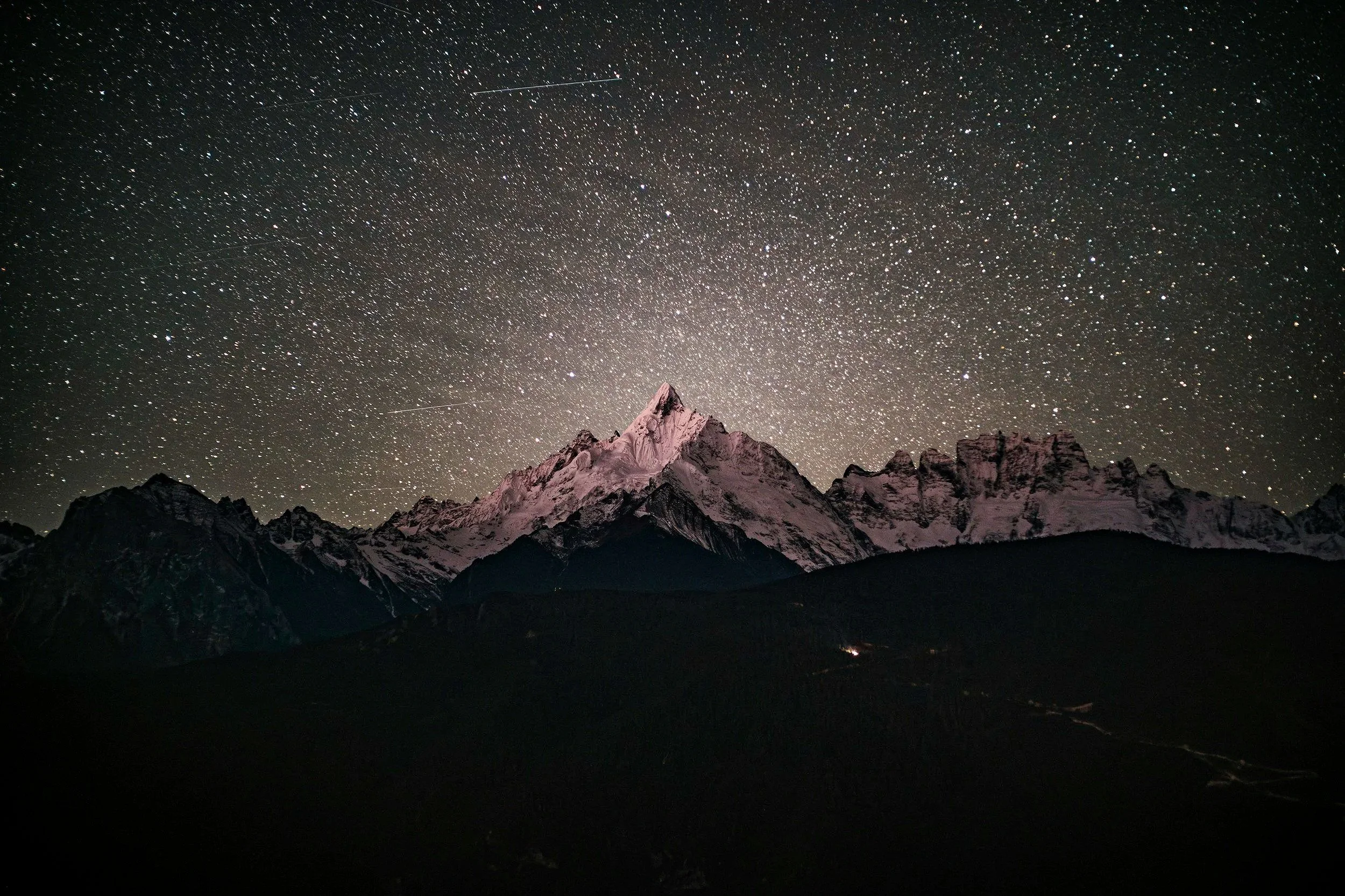 Nighttime view of snow-capped mountain peaks under a star-filled sky with visible shooting stars.