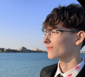 Close-up of a young person with short, wavy brown hair and glasses, standing outdoors near water under a clear blue sky.