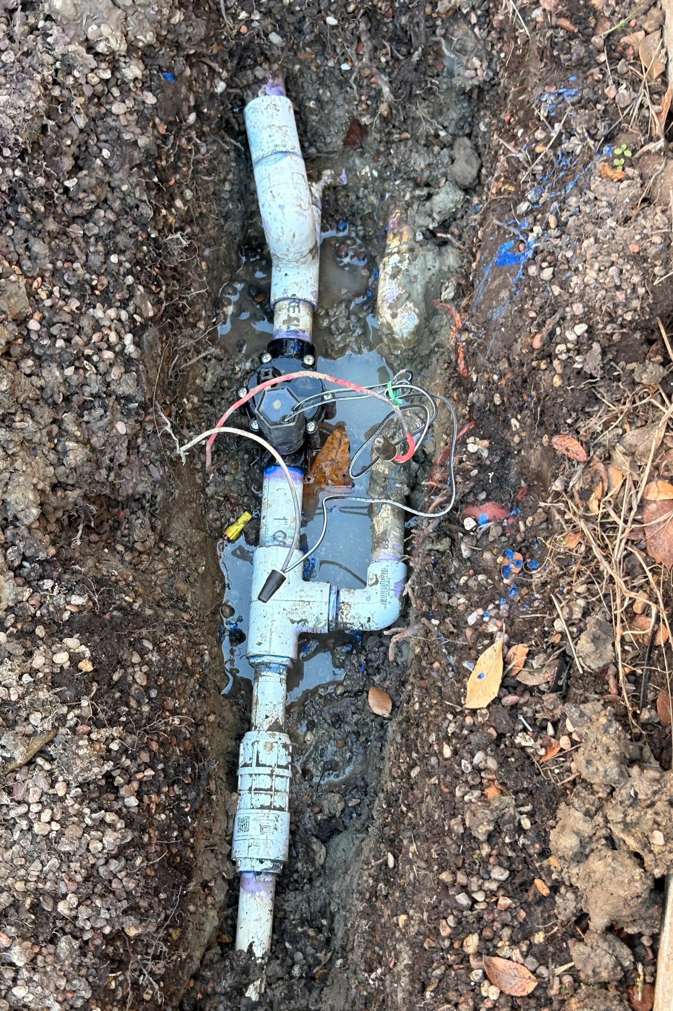 A damaged underground water pipe in a trench with soil and leaves around it.
