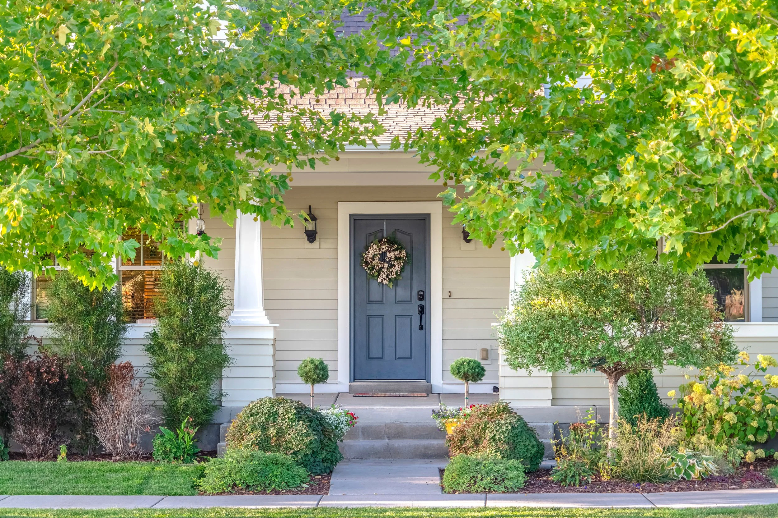 Front yard of a house with a dark gray door decorated with a flower wreath, surrounded by shrubs, trees, and green plants, under a leafy overhang.
