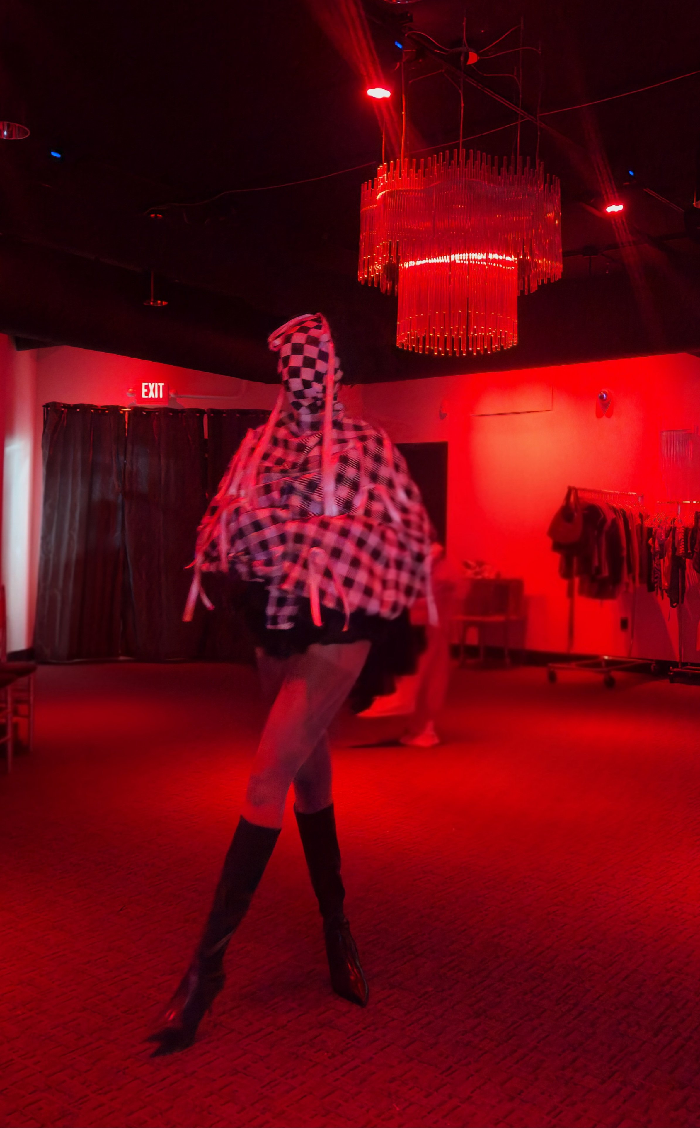 Person in a checkered hoodie and leggings dancing in a dark room with red lighting, a chandelier, and an exit sign.
