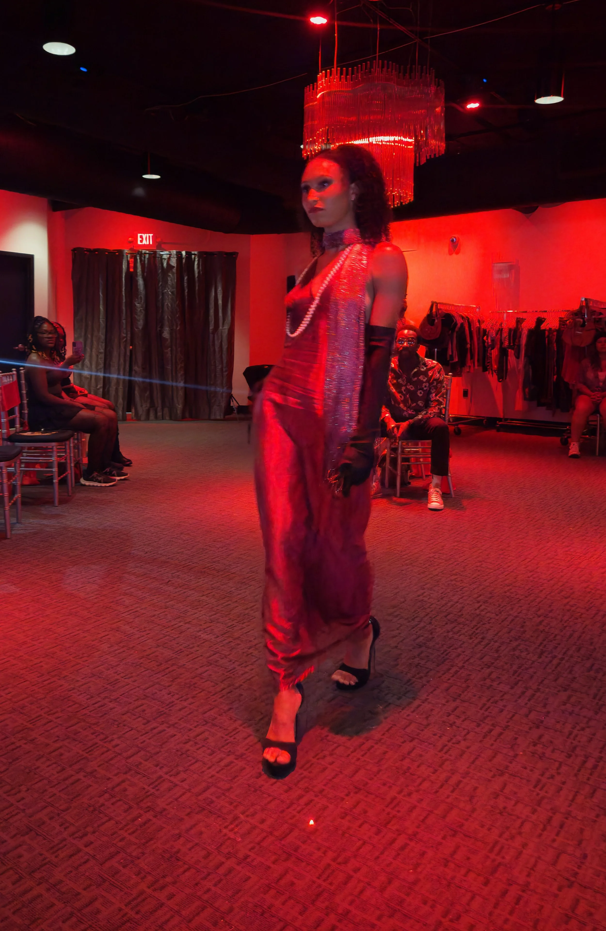 A runway show featuring a model walking on a red-lit stage, wearing a red satin dress with black gloves and high heels. Audience members are seated on either side, some taking pictures. The room has dark walls, clothing racks, and a large chandelier 