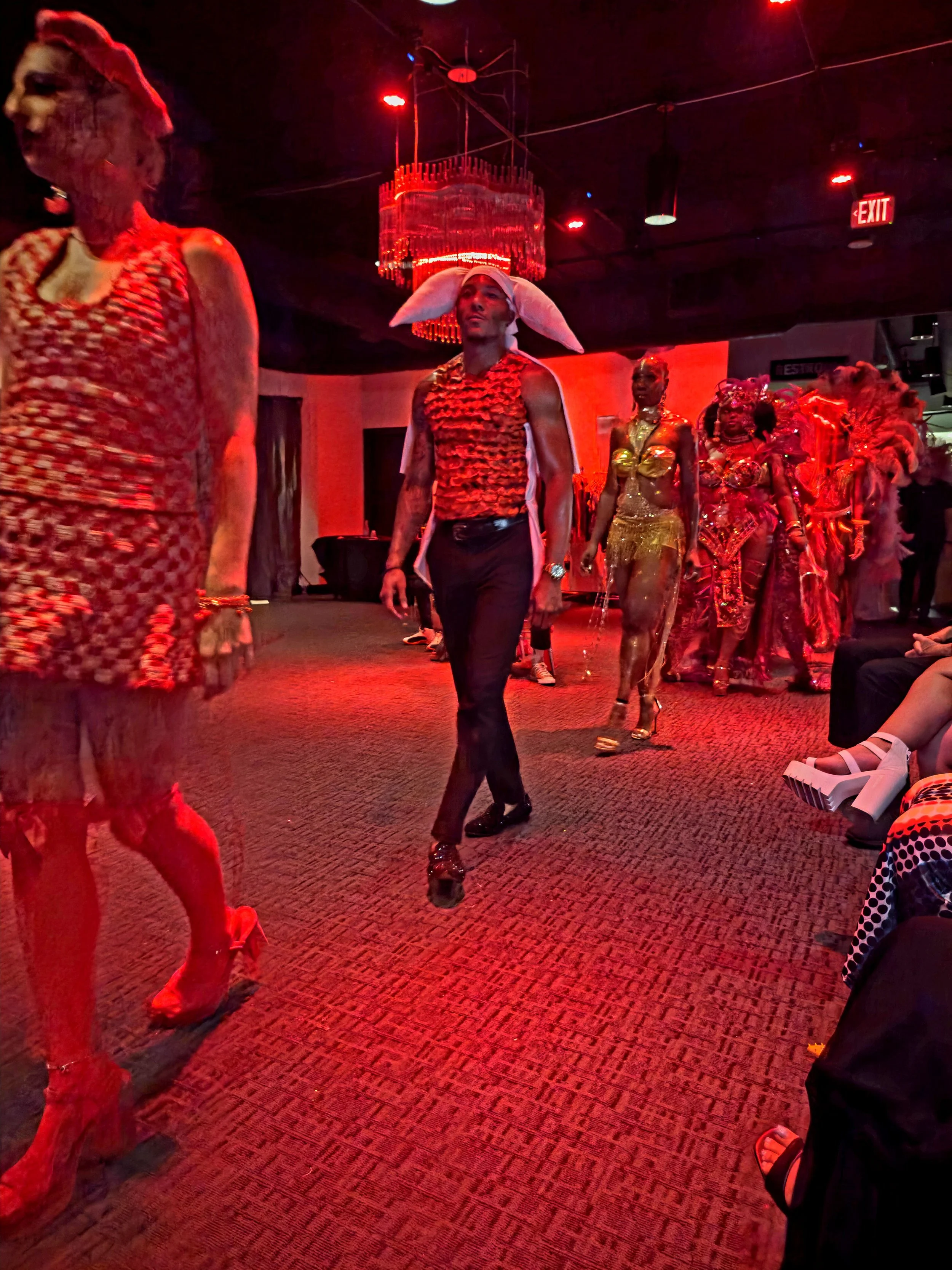 People dressed in colorful, elaborate costumes walking on stage under red lighting at a carnival or festival event.