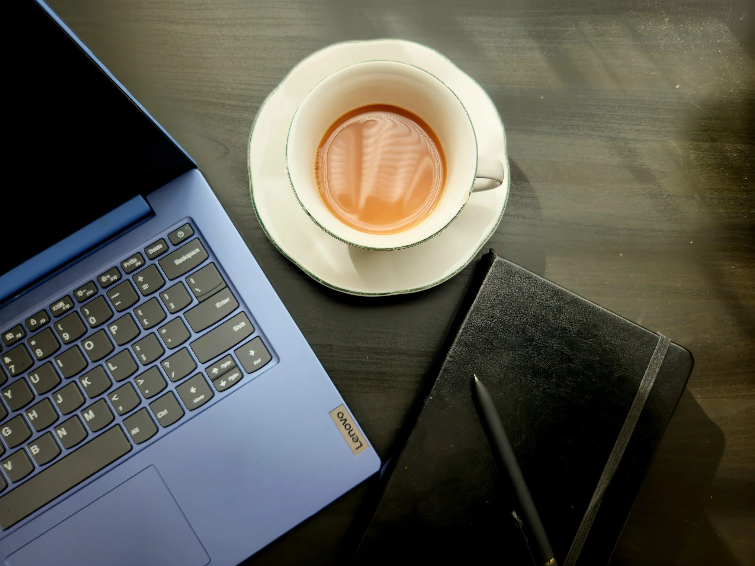 Overhead view of a workspace showing a laptop, a cup of tea on a saucer, a closed notebook, and a pen on a dark wooden surface.