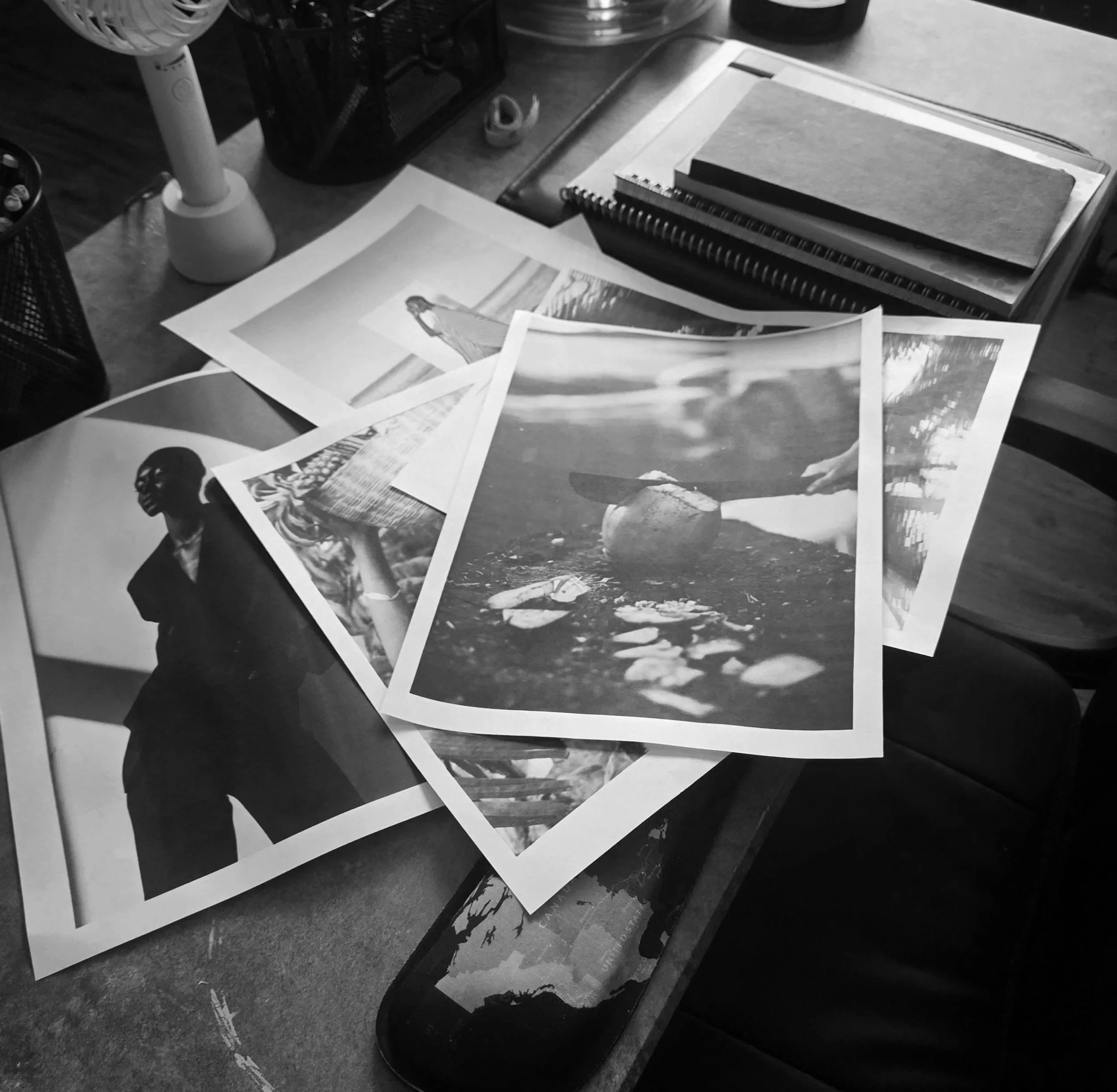 A cluttered desk with scattered photographs, a spiral notebook, a closed laptop, a small fan, and various office supplies on a dark surface.