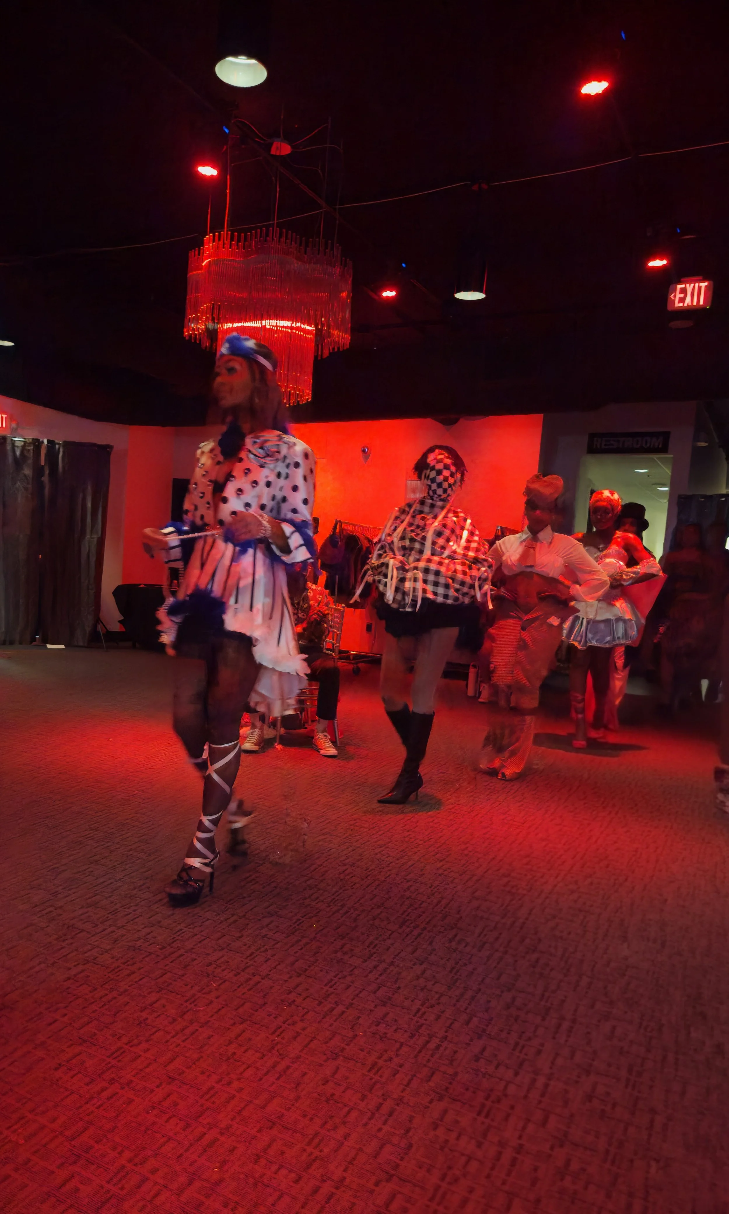 People dressed in colorful, theatrical costumes dancing under red lighting in a nightclub or performance venue.