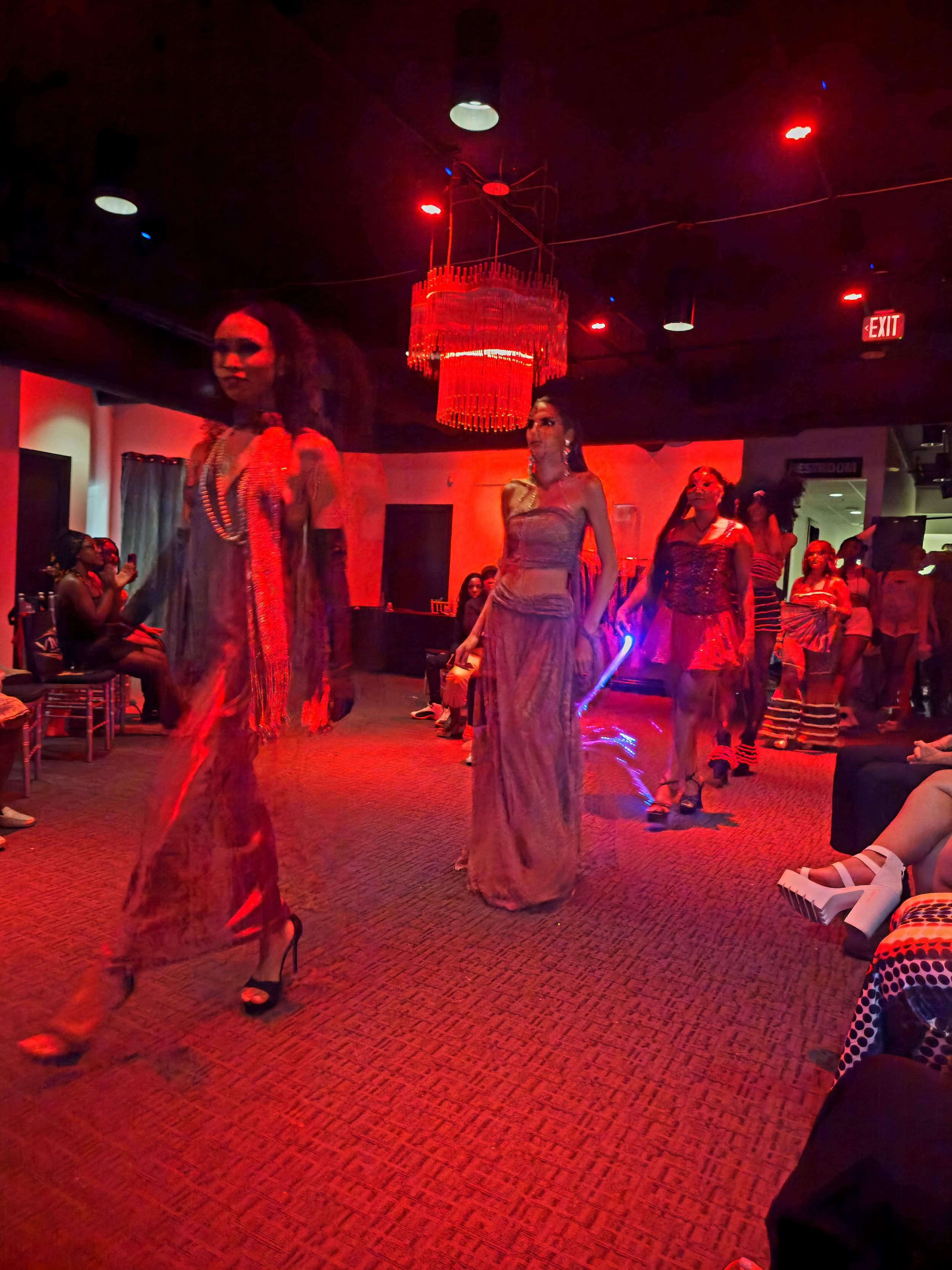 People dancing and socializing in a dimly lit nightclub with red lighting, a chandelier, and red exit sign.