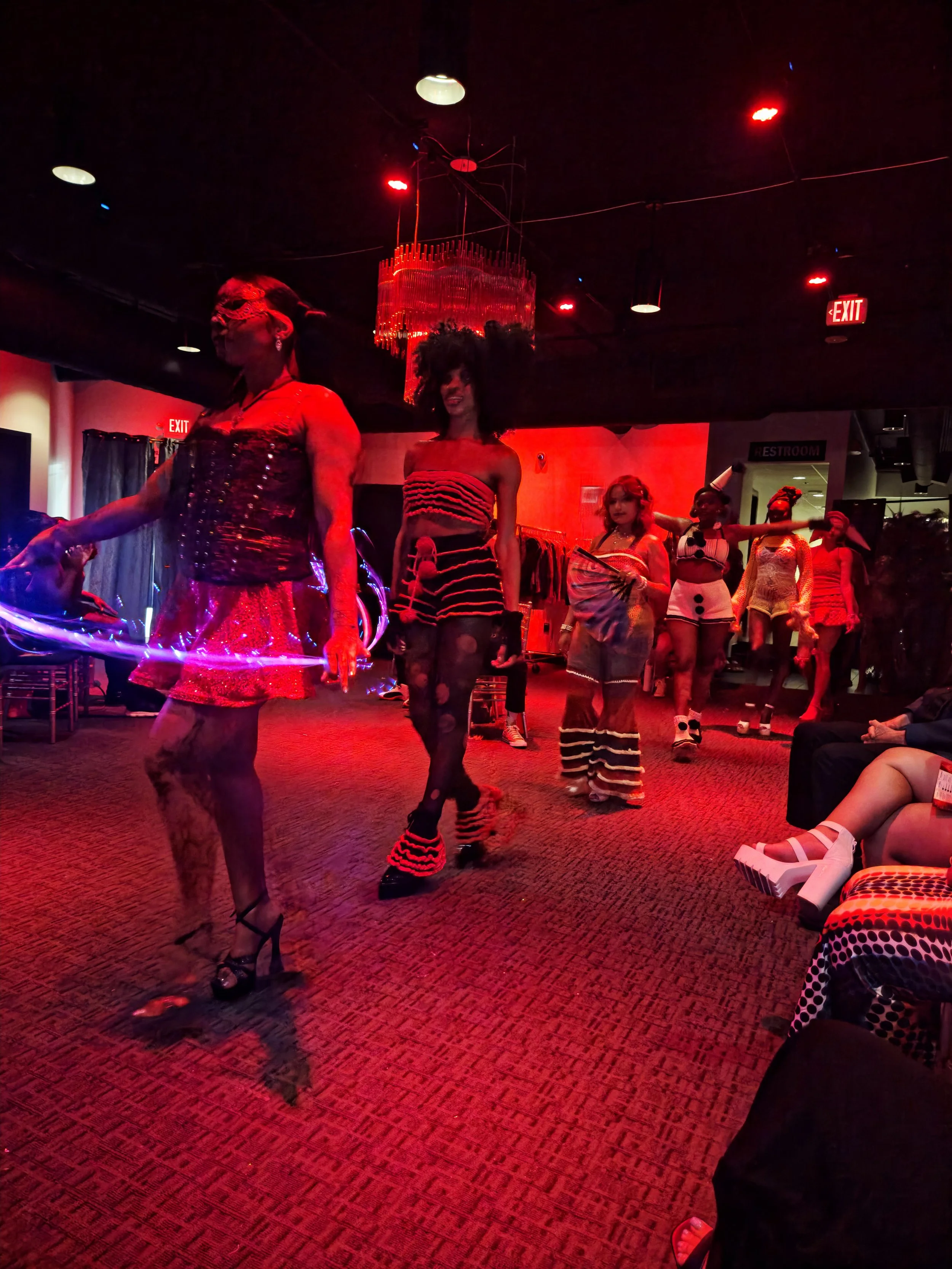 Performance scene featuring multiple individuals dressed in colorful, eclectic costumes, illuminated by vivid red and purple lighting, with audience members seated in the background.