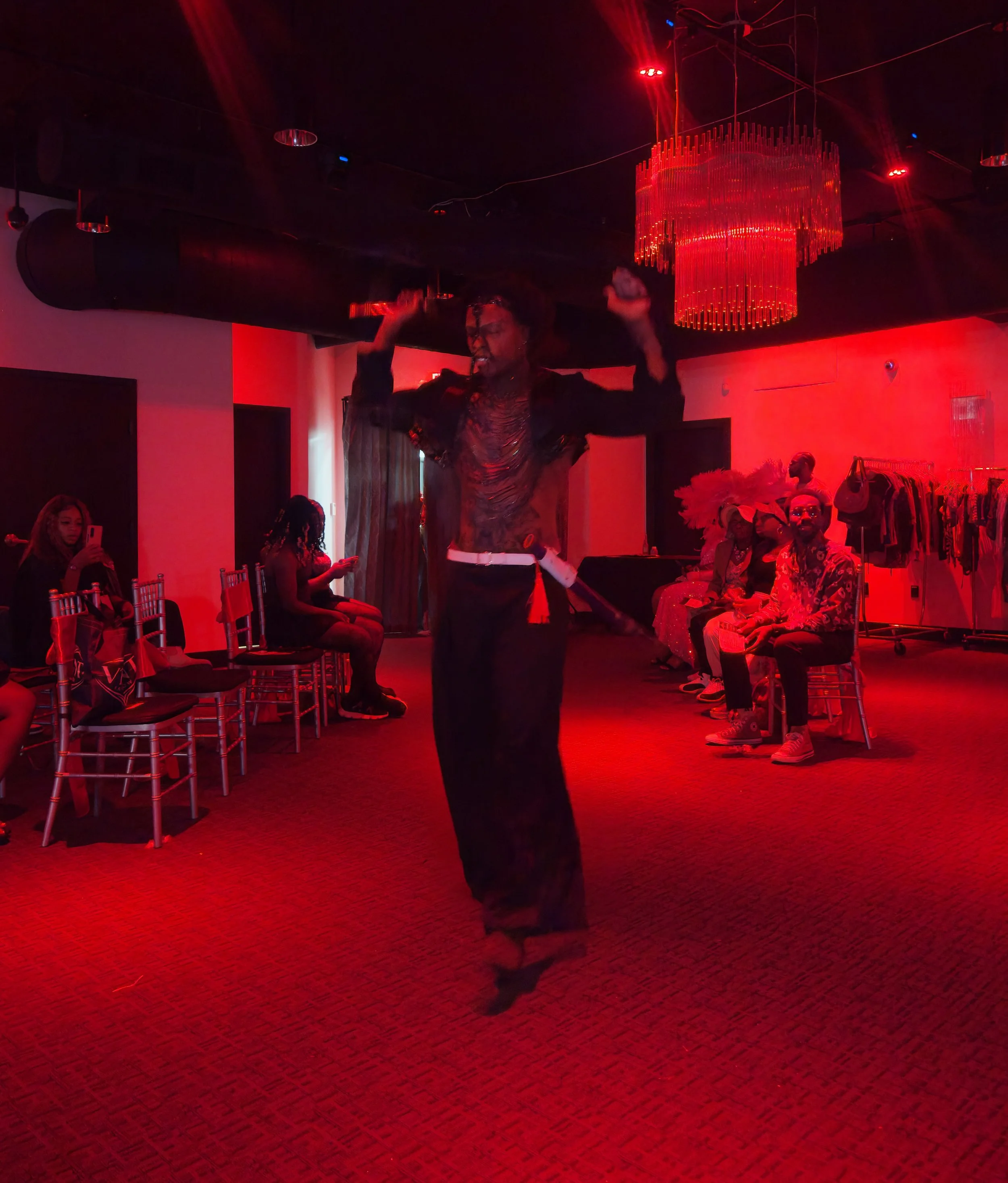 Person in black outfit dancing in a room with red lighting, surrounded by seated spectators and a red chandelier.