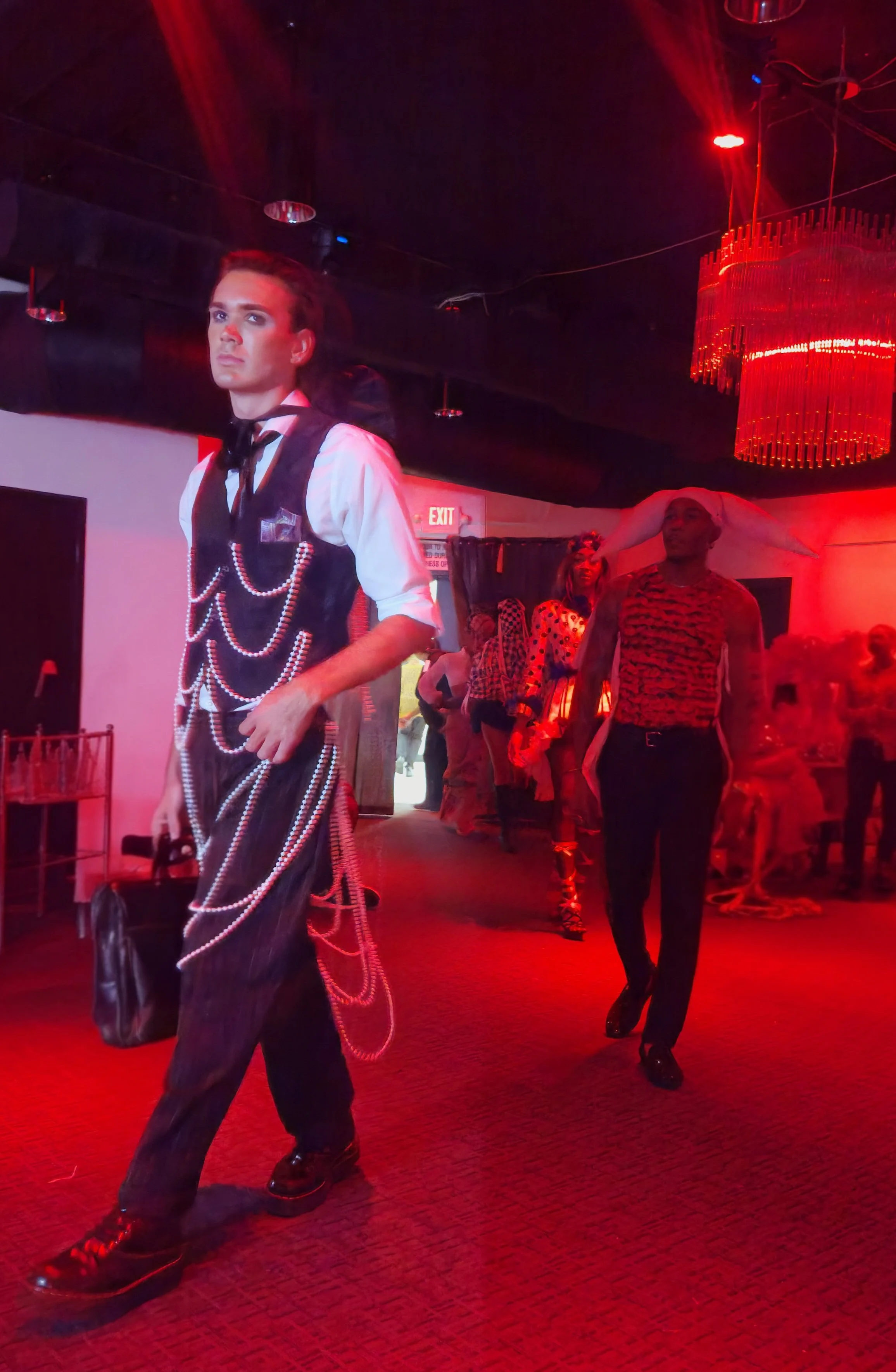 A person with a serious expression wearing a black vest decorated with pearl chains, black pants, and shiny shoes, walking in a dimly lit room with red lighting. Other people in colorful costumes, including one with a white hat with long white ears, 