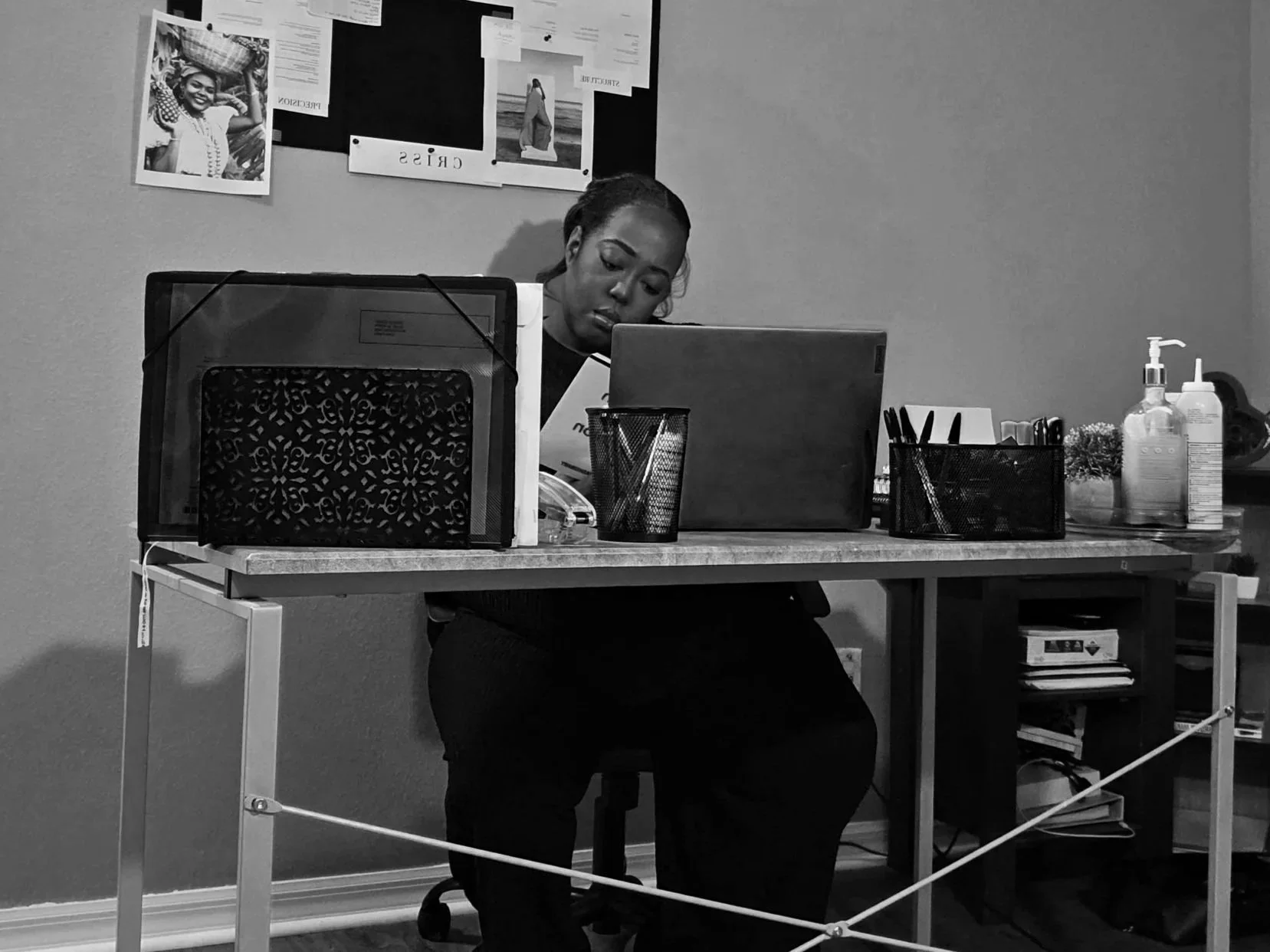 A woman sitting at a cluttered desk with a computer, papers, and office supplies, looking down at her work environment, in a room with a bulletin board on the wall.
