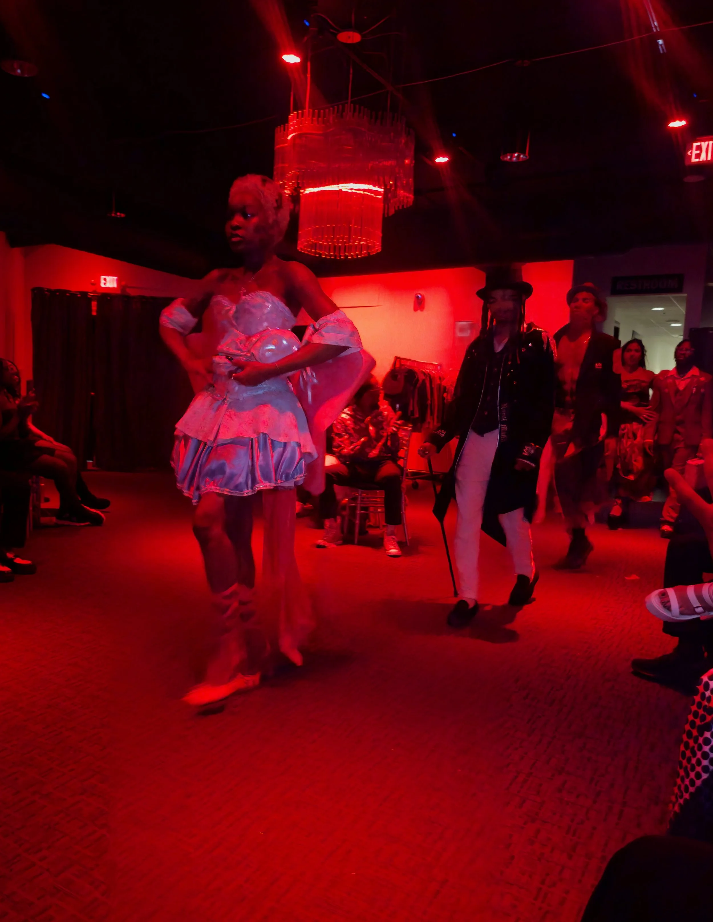 People dancing in a dimly lit club or party venue with red lighting and a chandelier on the ceiling.