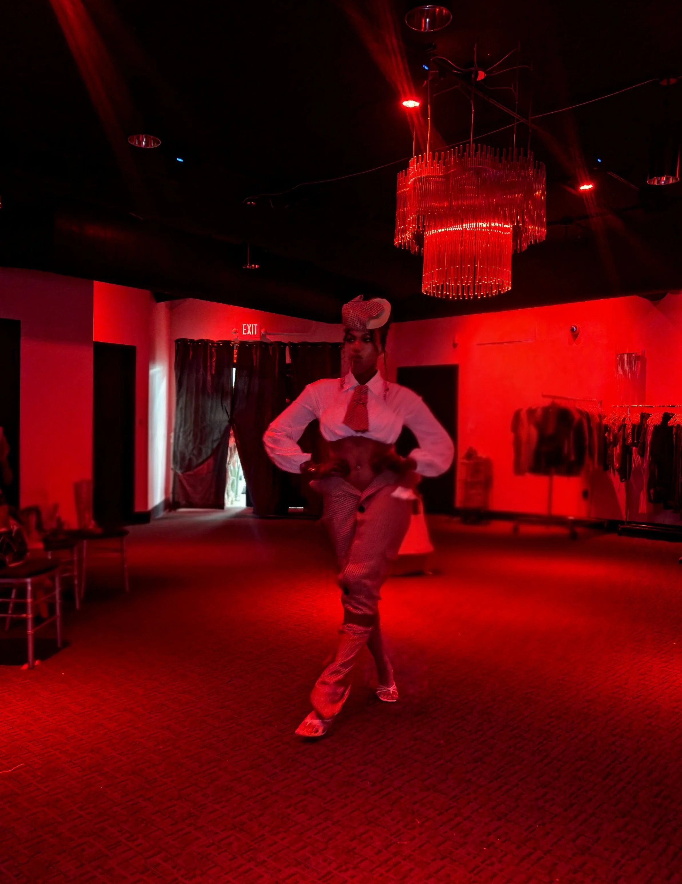 A person dancing alone in a dimly lit room illuminated by red light, with a large red chandelier hanging from the ceiling and clothing racks visible in the background.