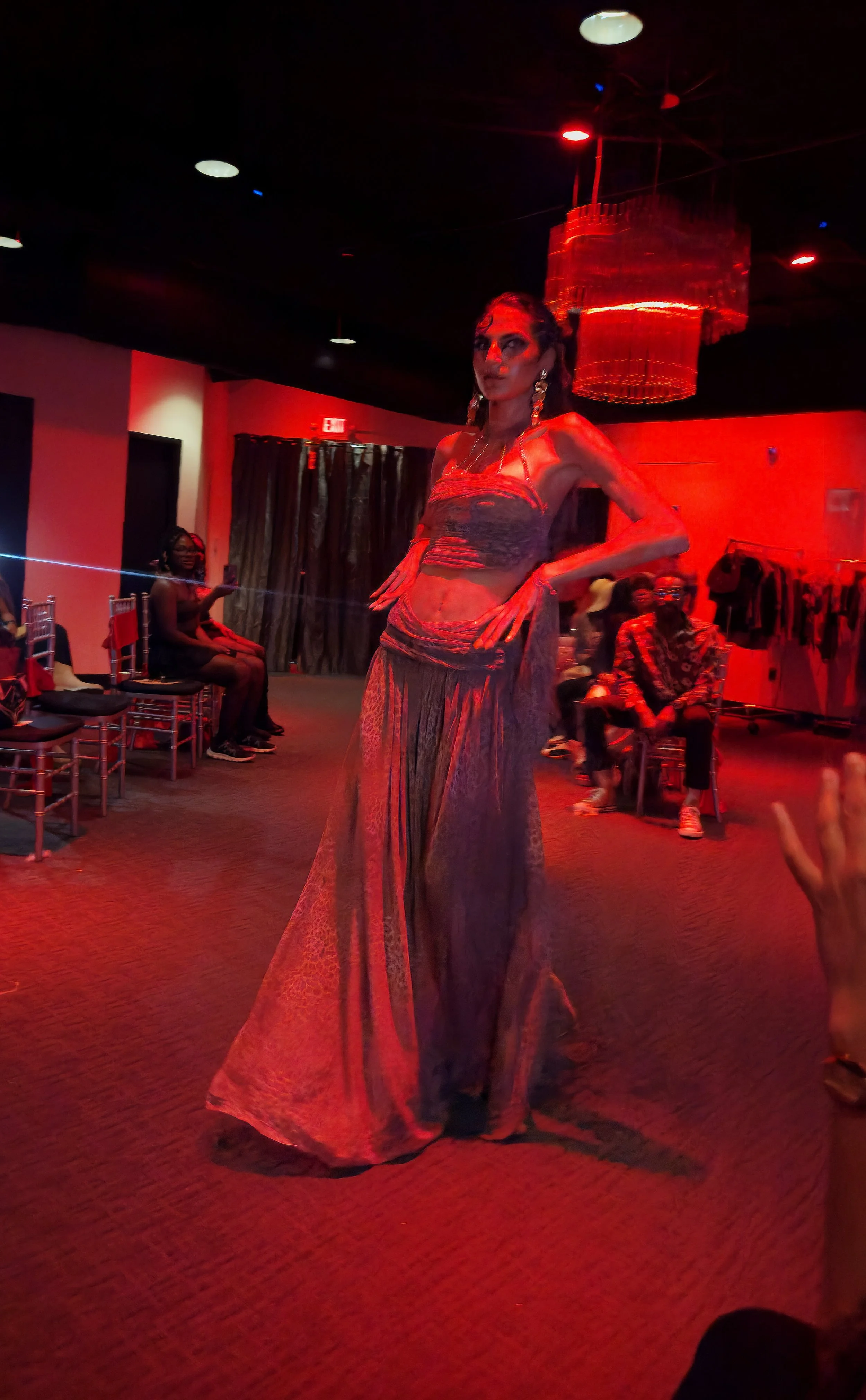 Drag performer on stage in a colorful costume, with audience members seated in the background, under red lighting.