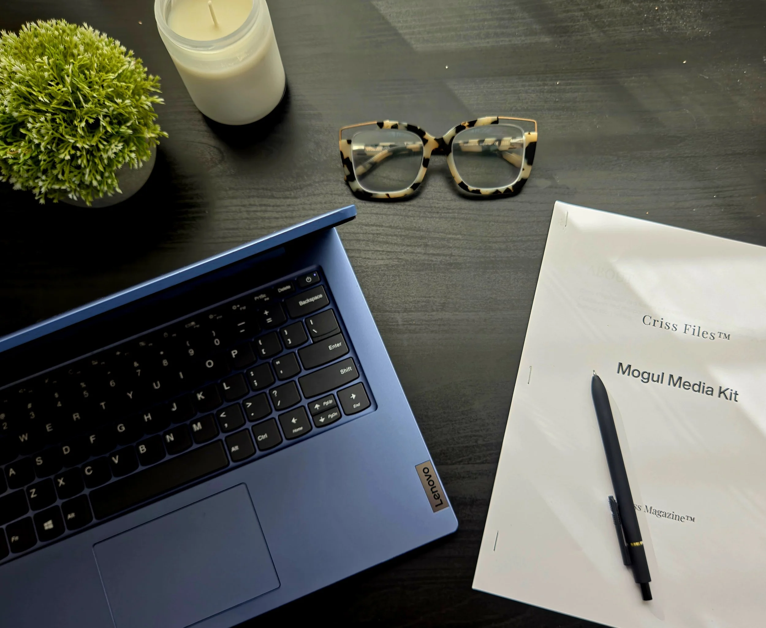 A workspace with a potted plant, a lit white candle, eyeglasses with a leopard print frame, a closed white notebook, a black pen, a partially visible laptop, and a printed document on a wooden desk.