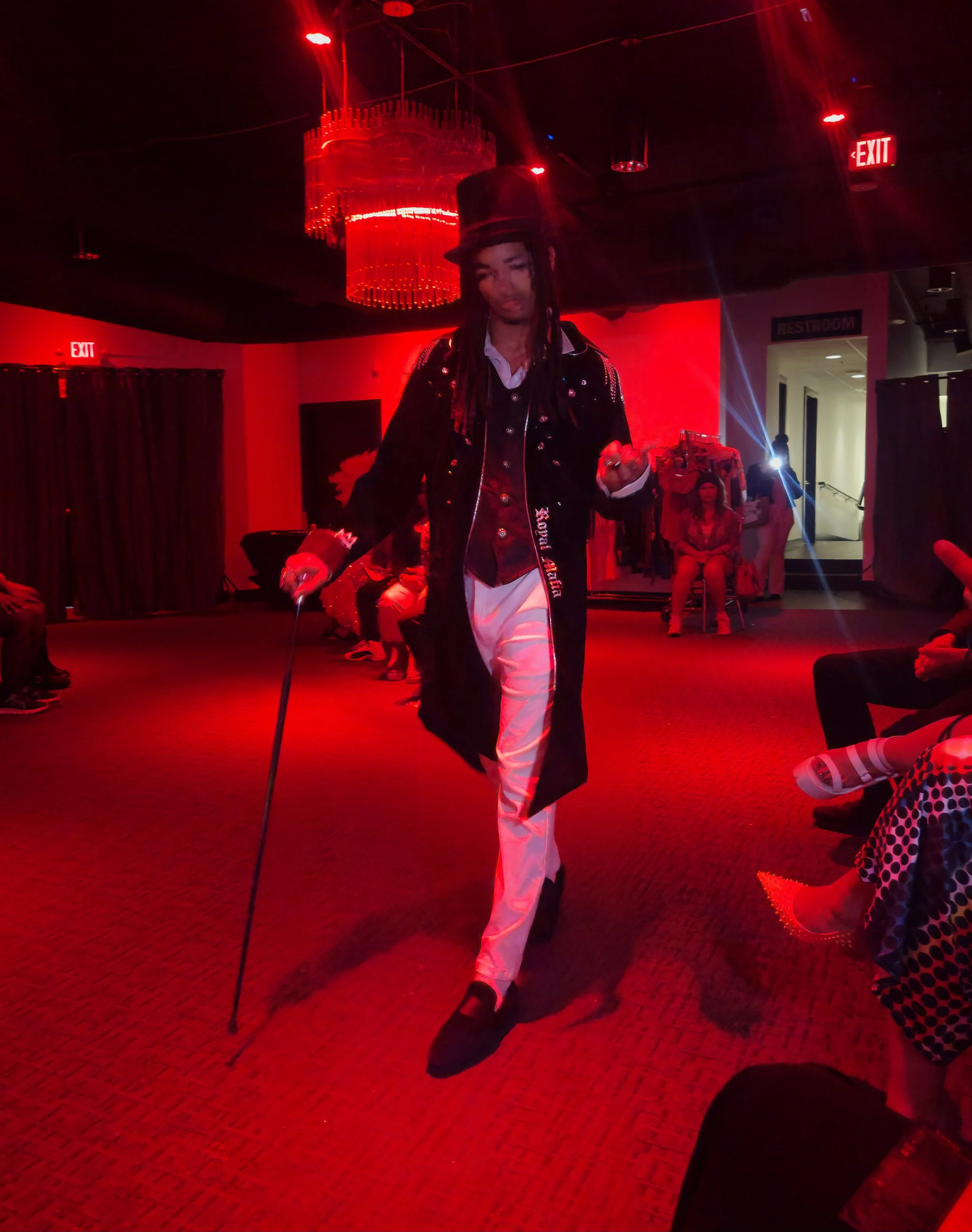 Person dressed in special costume, walking in a dimly lit room with red lighting, possibly dancing or performing, surrounded by seated audience members.