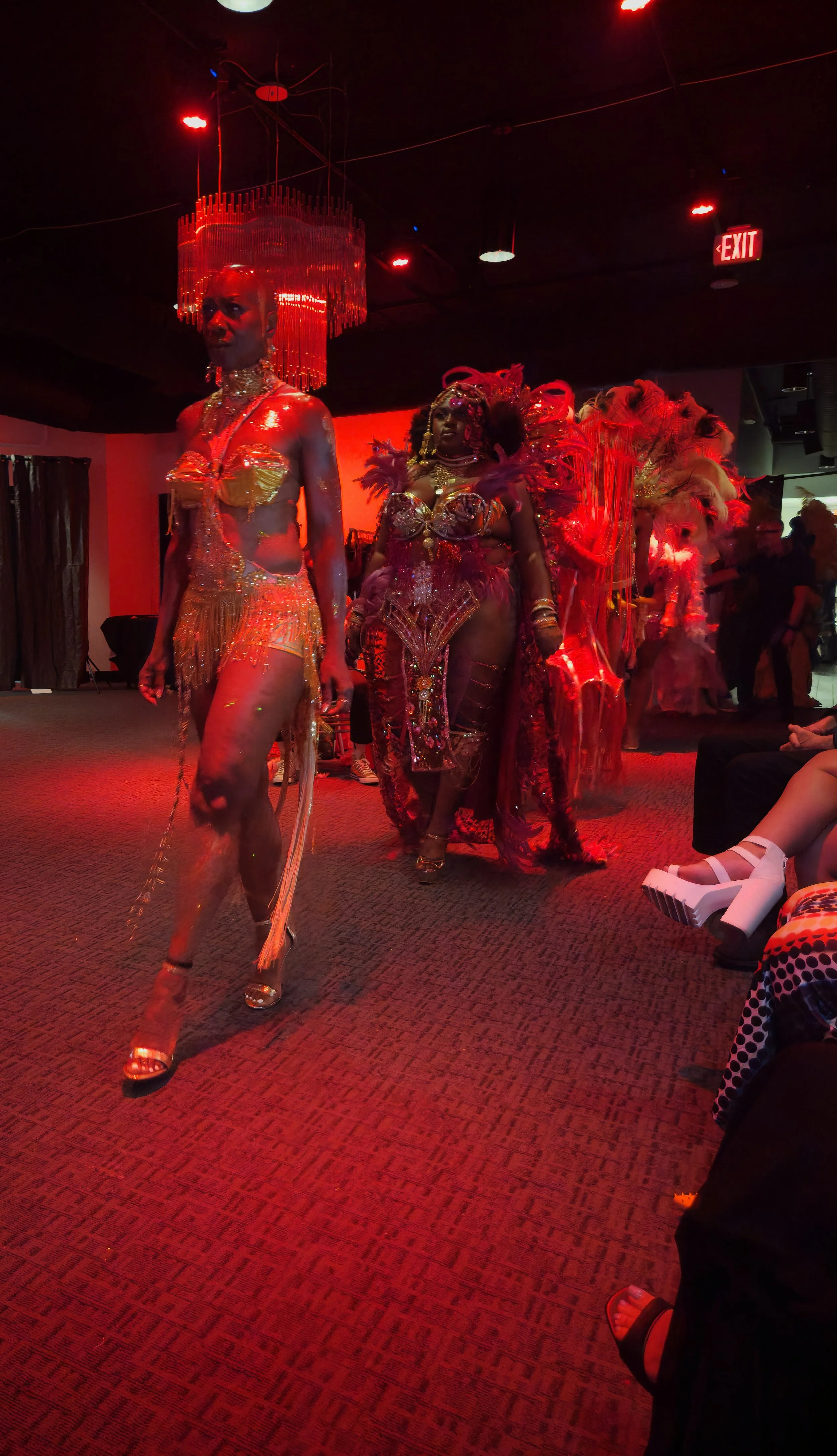 Performance with performers in elaborate, colorful costumes with feathers and sequins, walking in a line under red lighting in an indoor venue with a chandelier and exit sign visible.