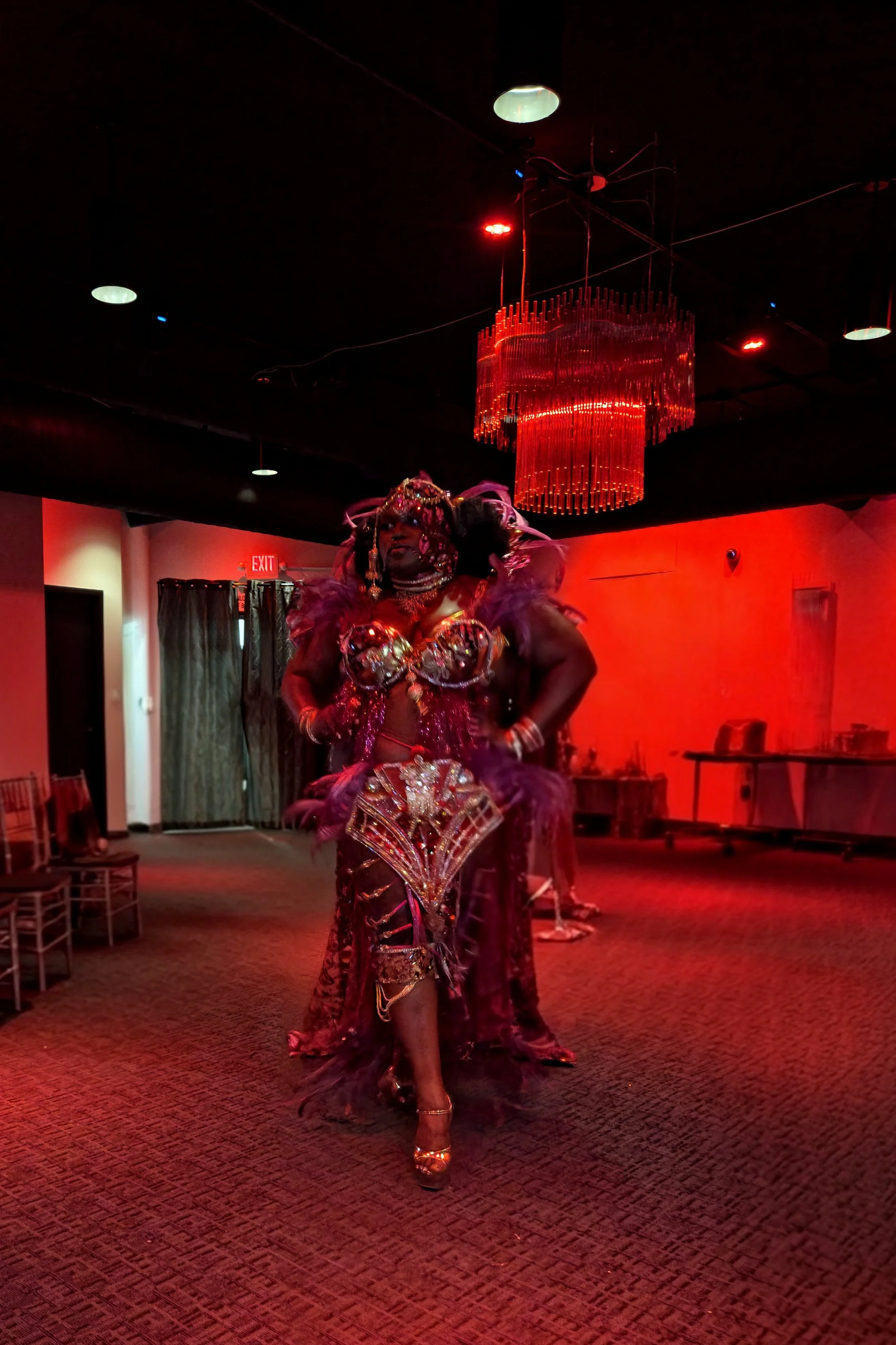A performer dressed in an elaborate, colorful costume with feathers, jewelry, and high-heeled shoes is standing in a red-lit room, possibly at a nightclub or performance space. The background includes a chandelier, exit sign, curtains, and chairs.