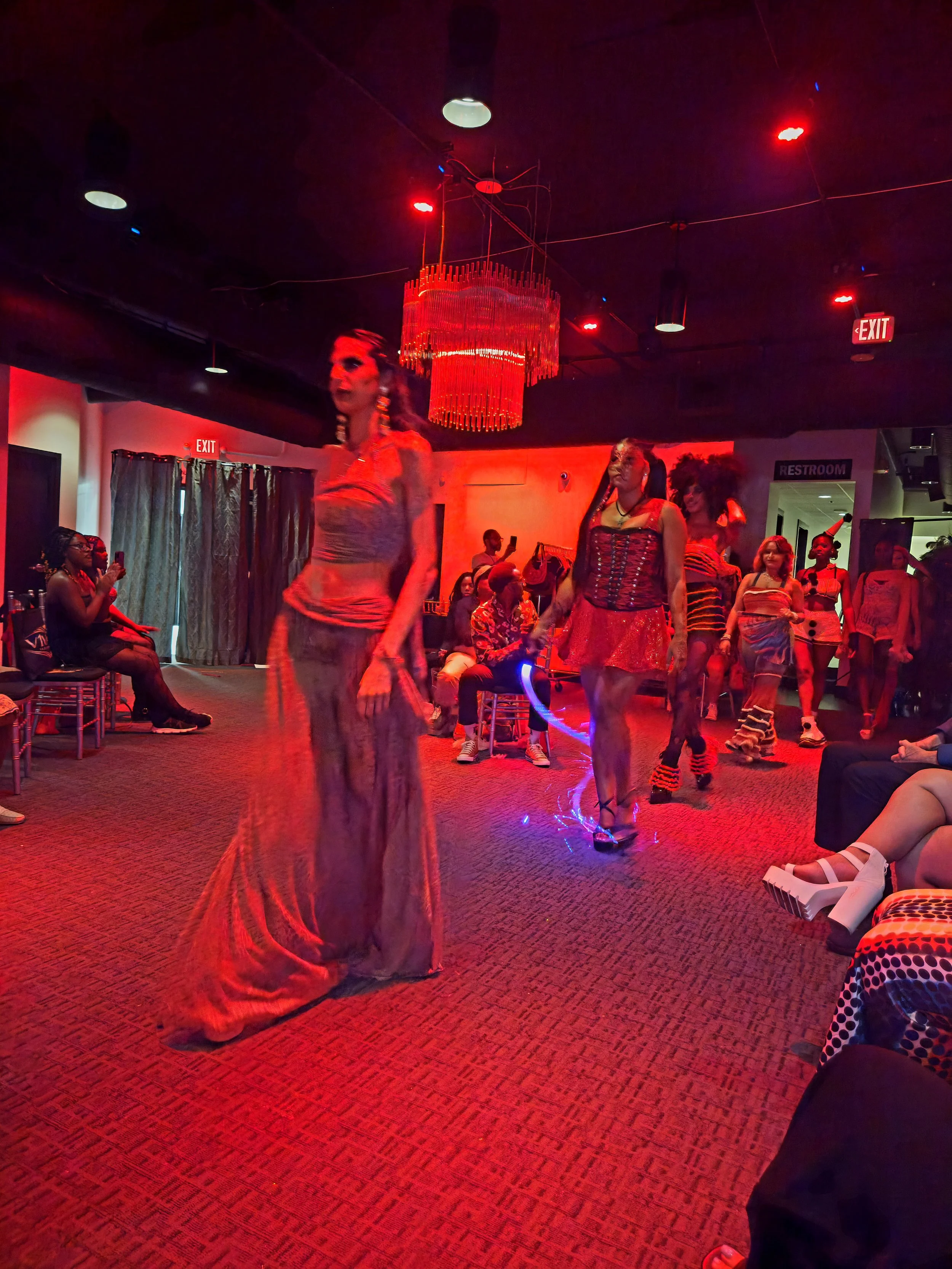 Group of performers and audience in a dimly lit room with red lighting, some performers wearing colorful costumes, including a woman in a red and black dress, a woman with long braids in a silver dress, and others in various costumes. A large red cha