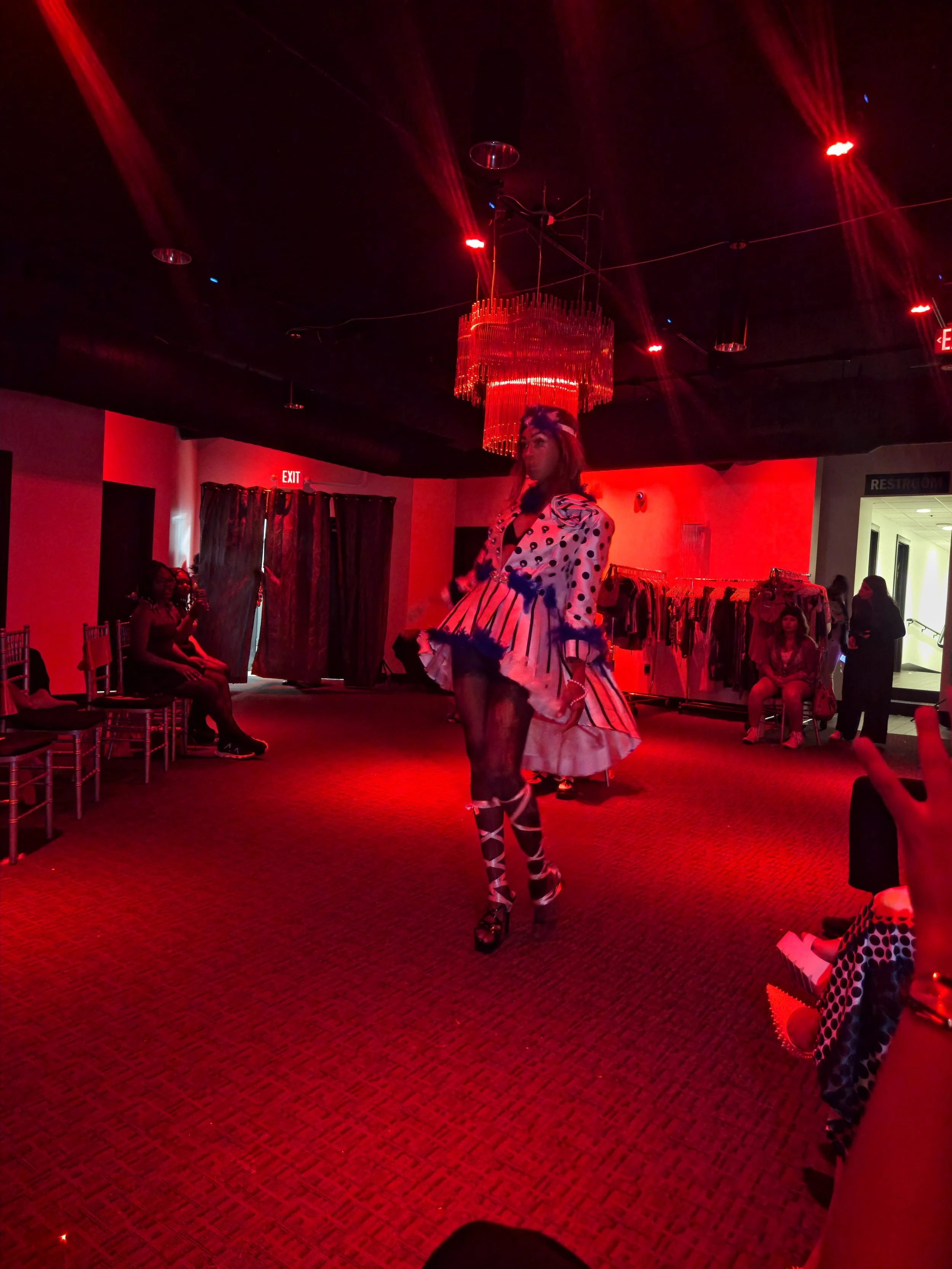 A woman in a polka dot dress with matching thigh-high boots walking in a room with red lighting and a chandelier hanging from the ceiling, with several people seated and standing.
