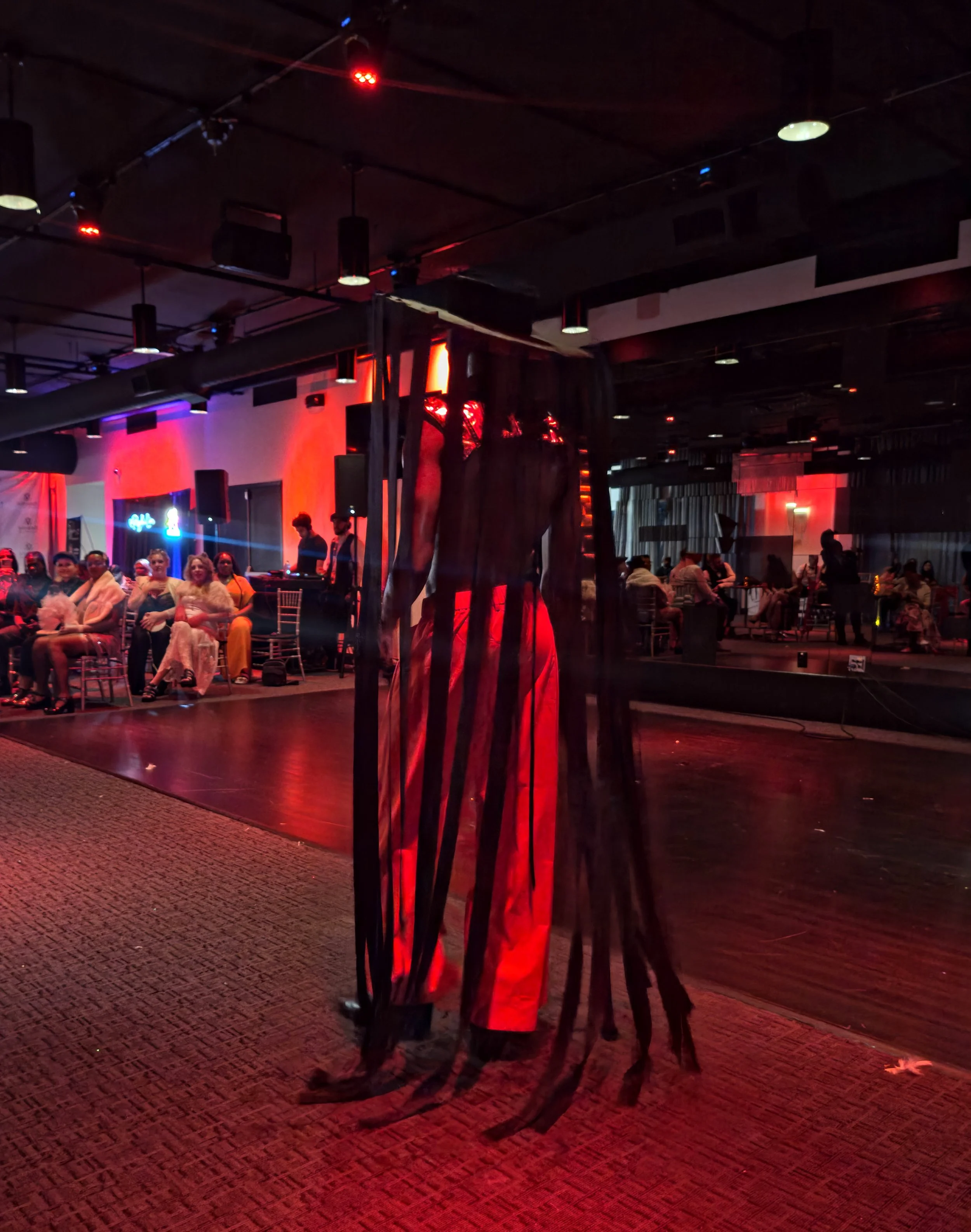 A performer wearing a long, flowing red and black striped cape dances on stage in a dimly lit venue with red lighting, while an audience watches from chairs.