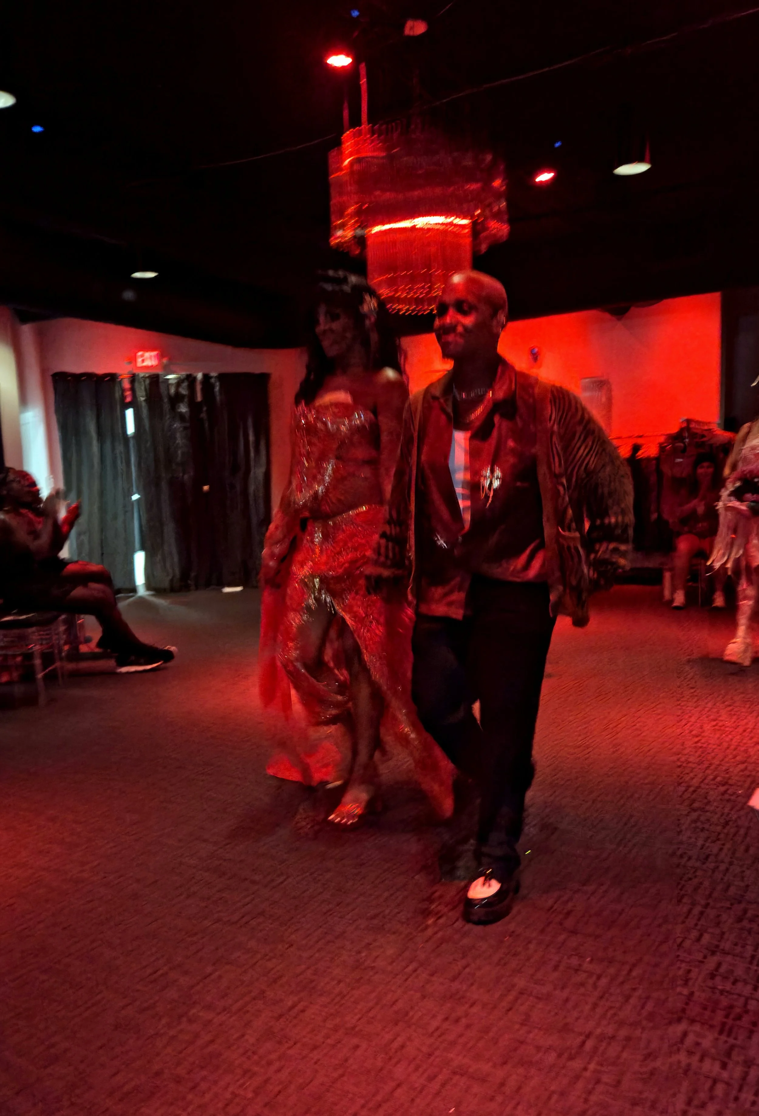 Two people dancing in a dimly lit room with red lighting, one in a shiny, colorful dress and the other in a patterned jacket and black pants.