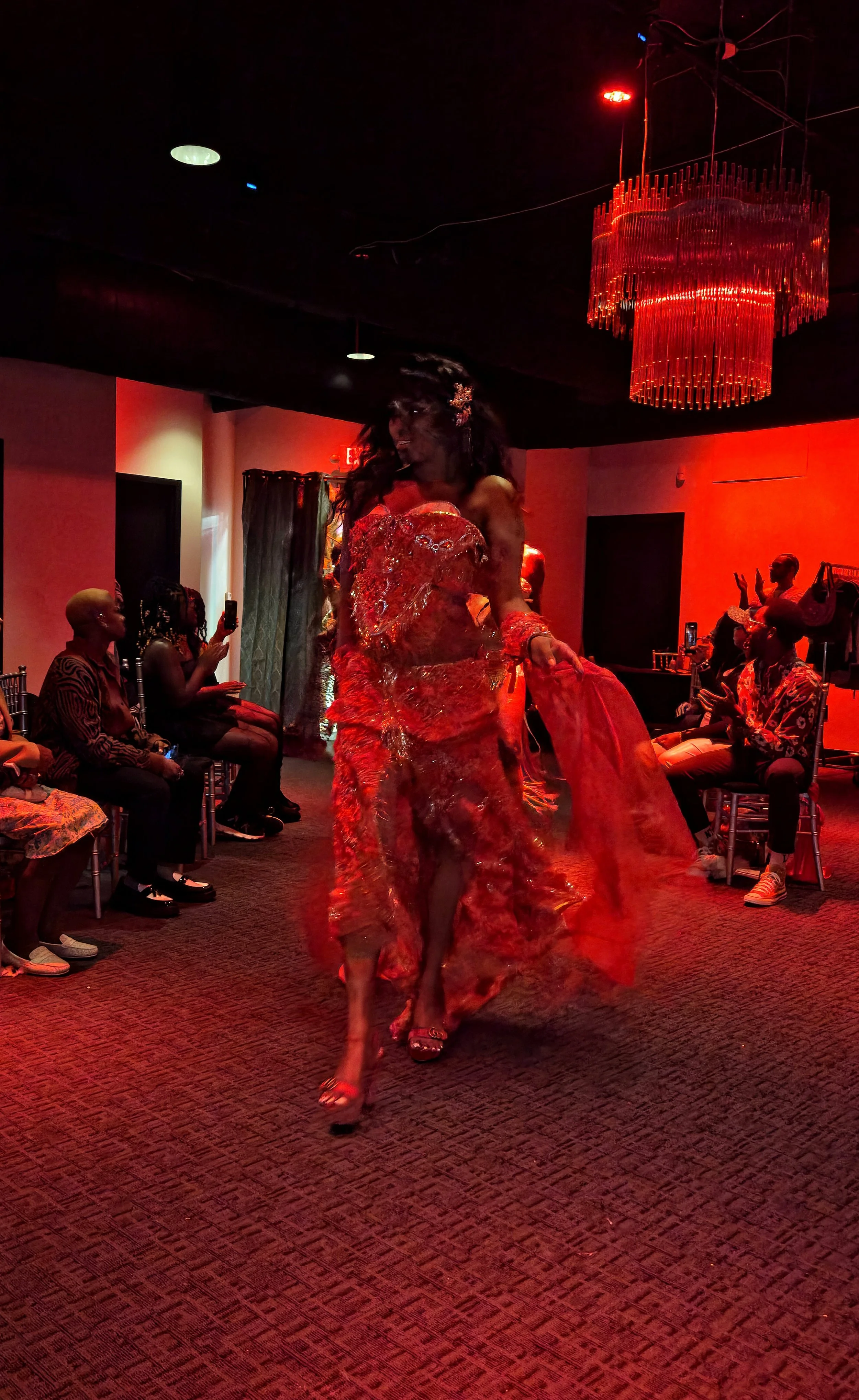A woman in a red, shiny gown walking on a runway at an indoor event with red lighting. The audience is seated along the sides, some taking photos.