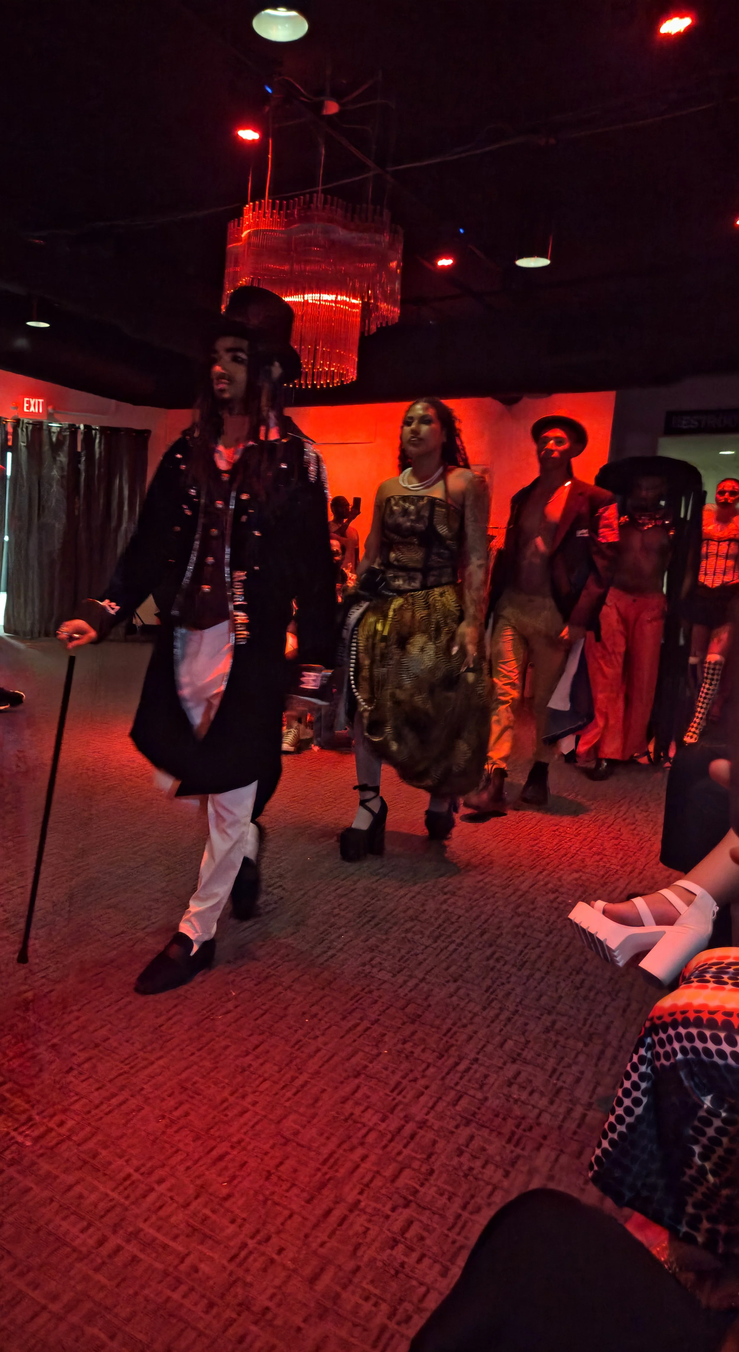 People walking in a nightclub with red lighting, some in elaborate costumes including a person with a top hat and cane, others in fashionable outfits, and a large red chandelier hanging from the ceiling.
