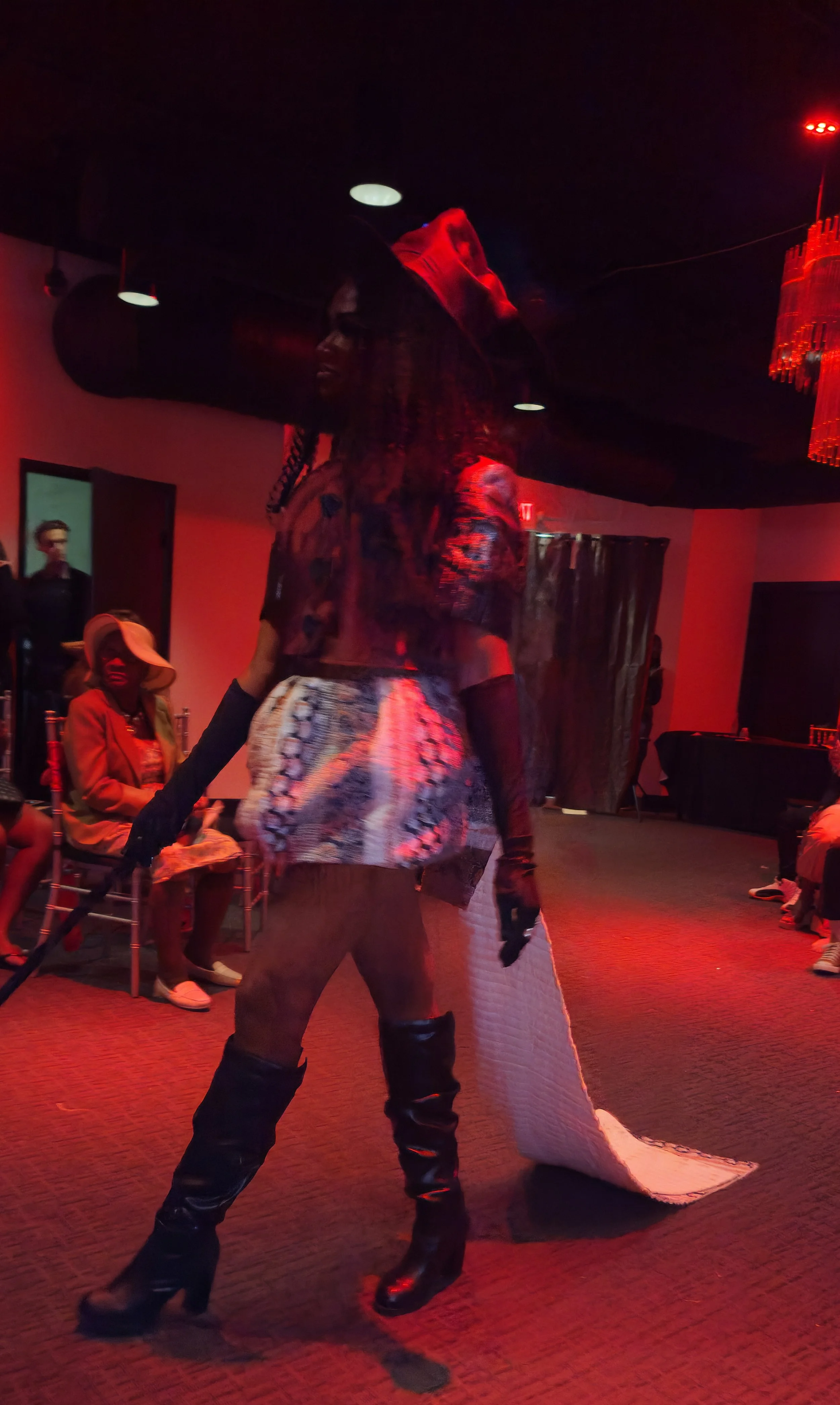 Person walking in a dimly lit room with red lighting, wearing black high-heeled boots, patterned shorts, and a dark top, with a white quilted blanket trailing behind. Several seated people are visible in the background, some watching and some smiling.