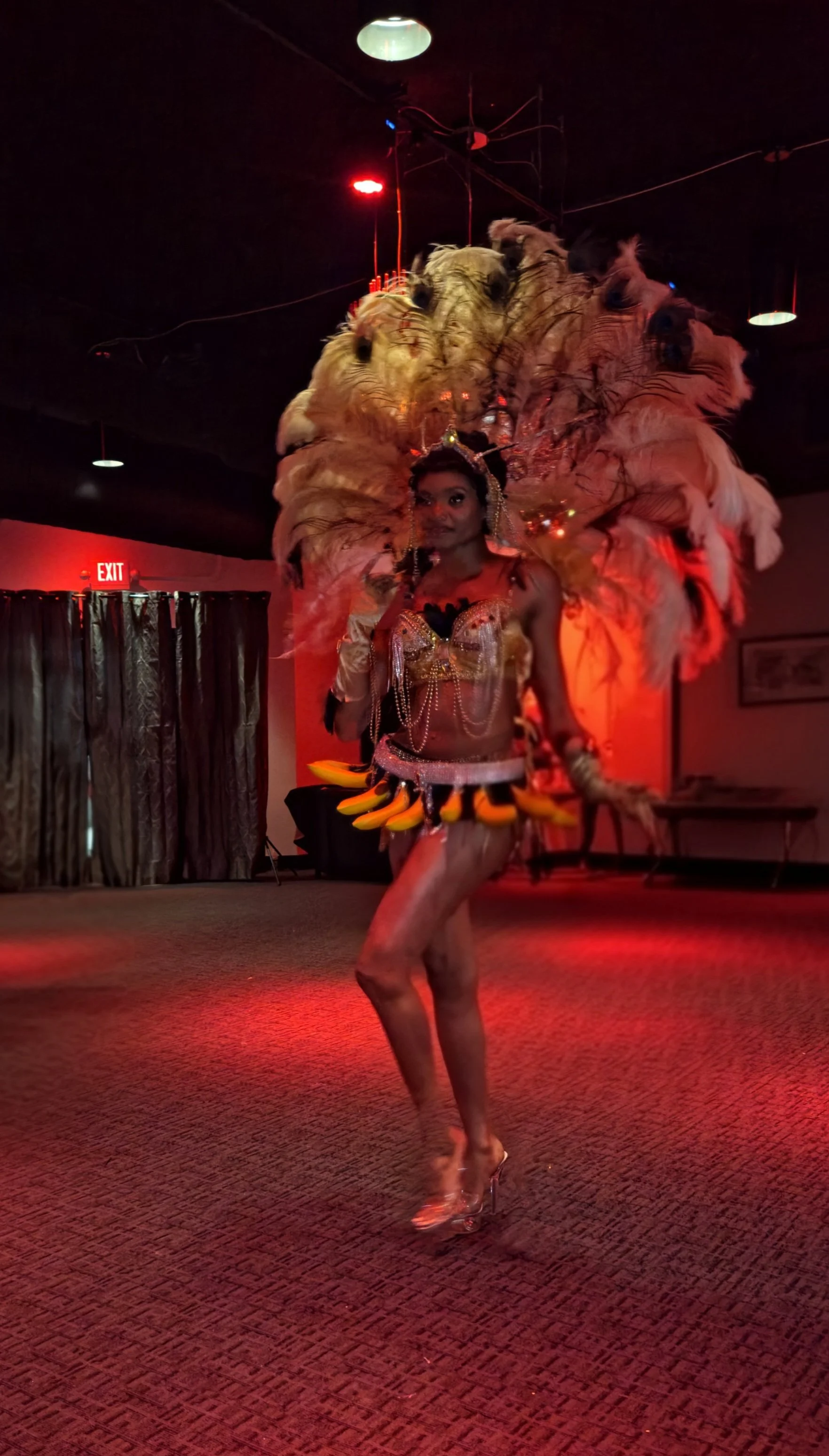 A performer in an elaborate costume with large feathered headpiece and a small skirt decorated with bananas, dancing in a dimly lit room with red lighting.