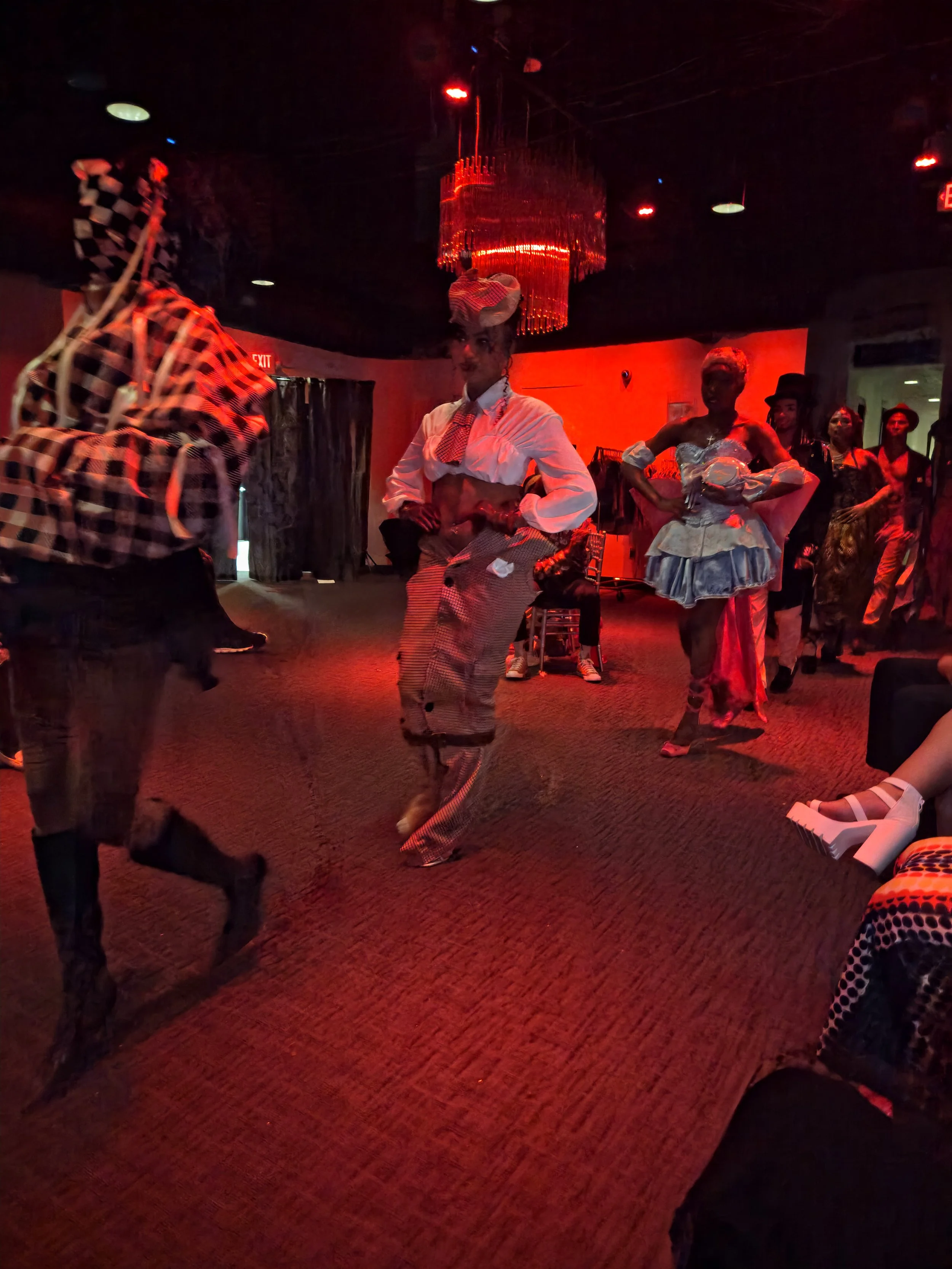 People dressed in vintage and theatrical costumes dancing and socializing in a dimly lit lounge with red lighting, chandelier, and patterned carpet.